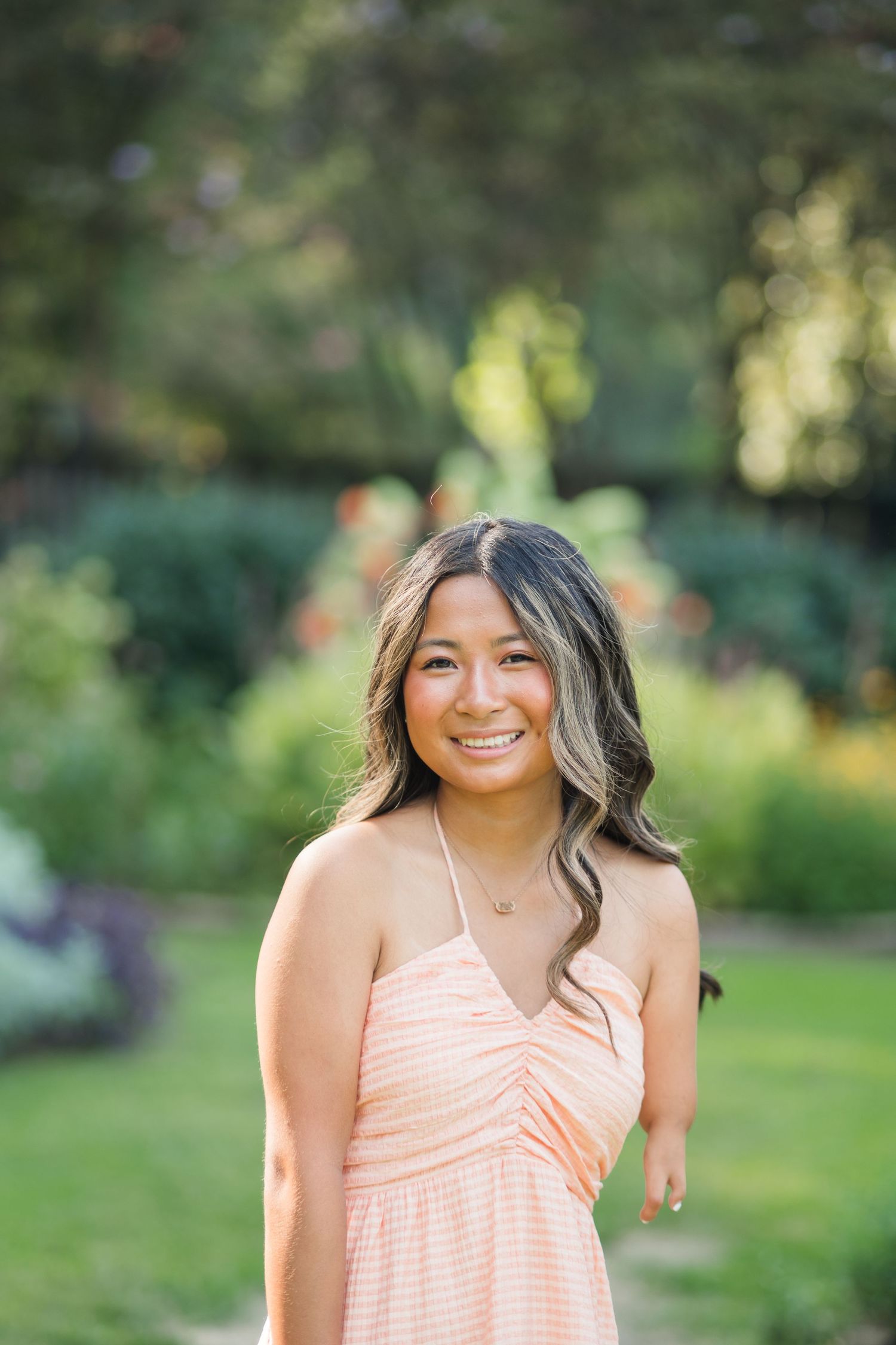 Young woman in a peach sundress smiling outdoors in a lush garden setting on a sunny day.