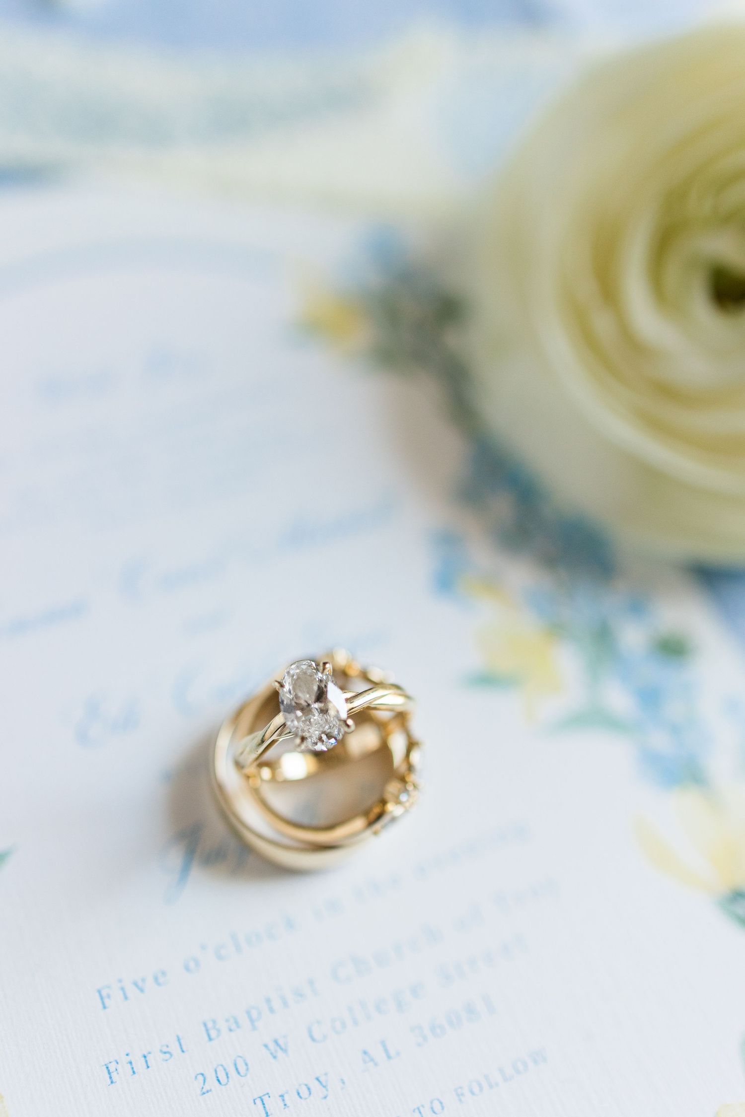 Gold wedding rings and diamond engagement ring stacked on a floral wedding invitation with a white ranunculus.