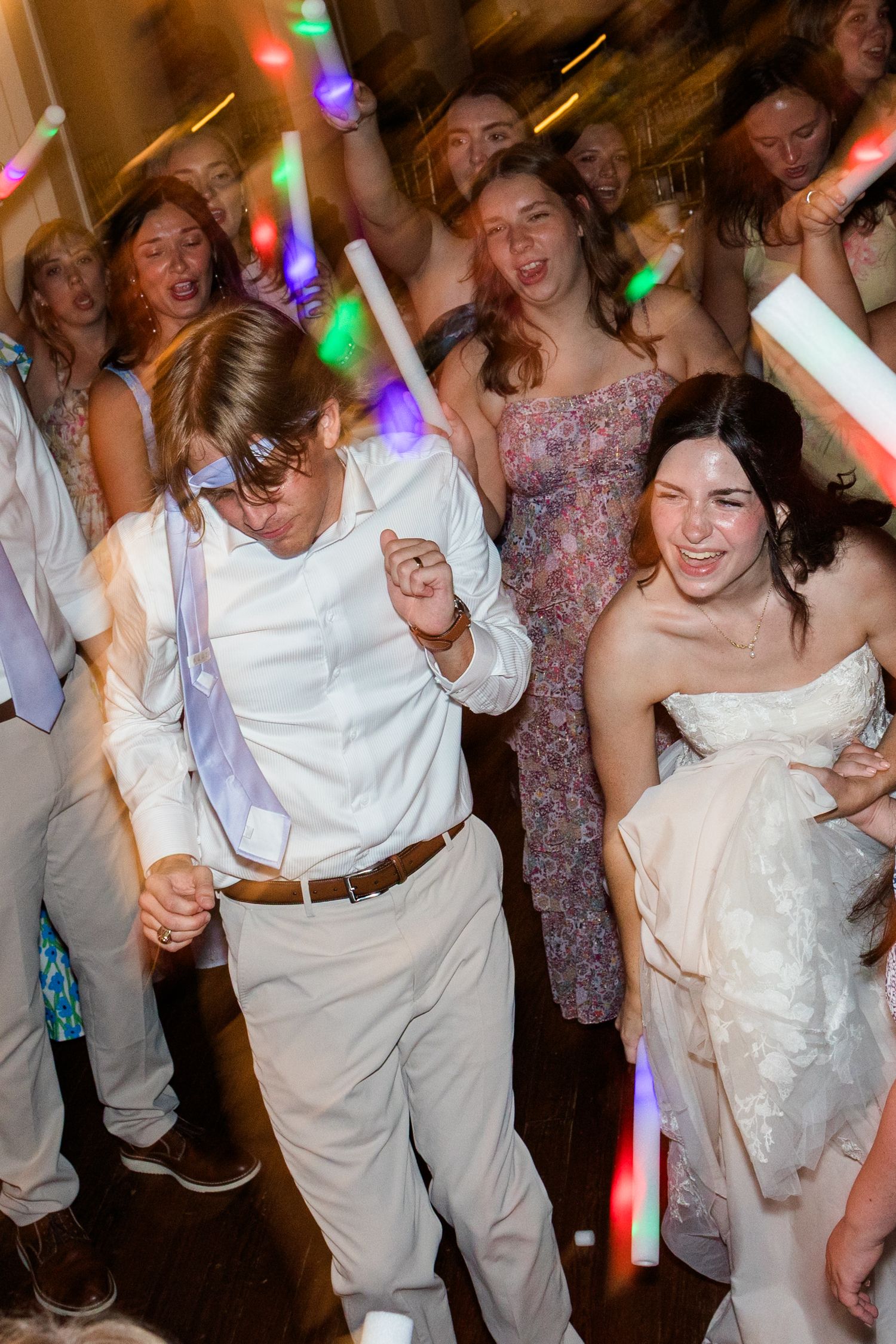 Bride and groom dancing joyfully with guests holding glow sticks at lively wedding reception party.