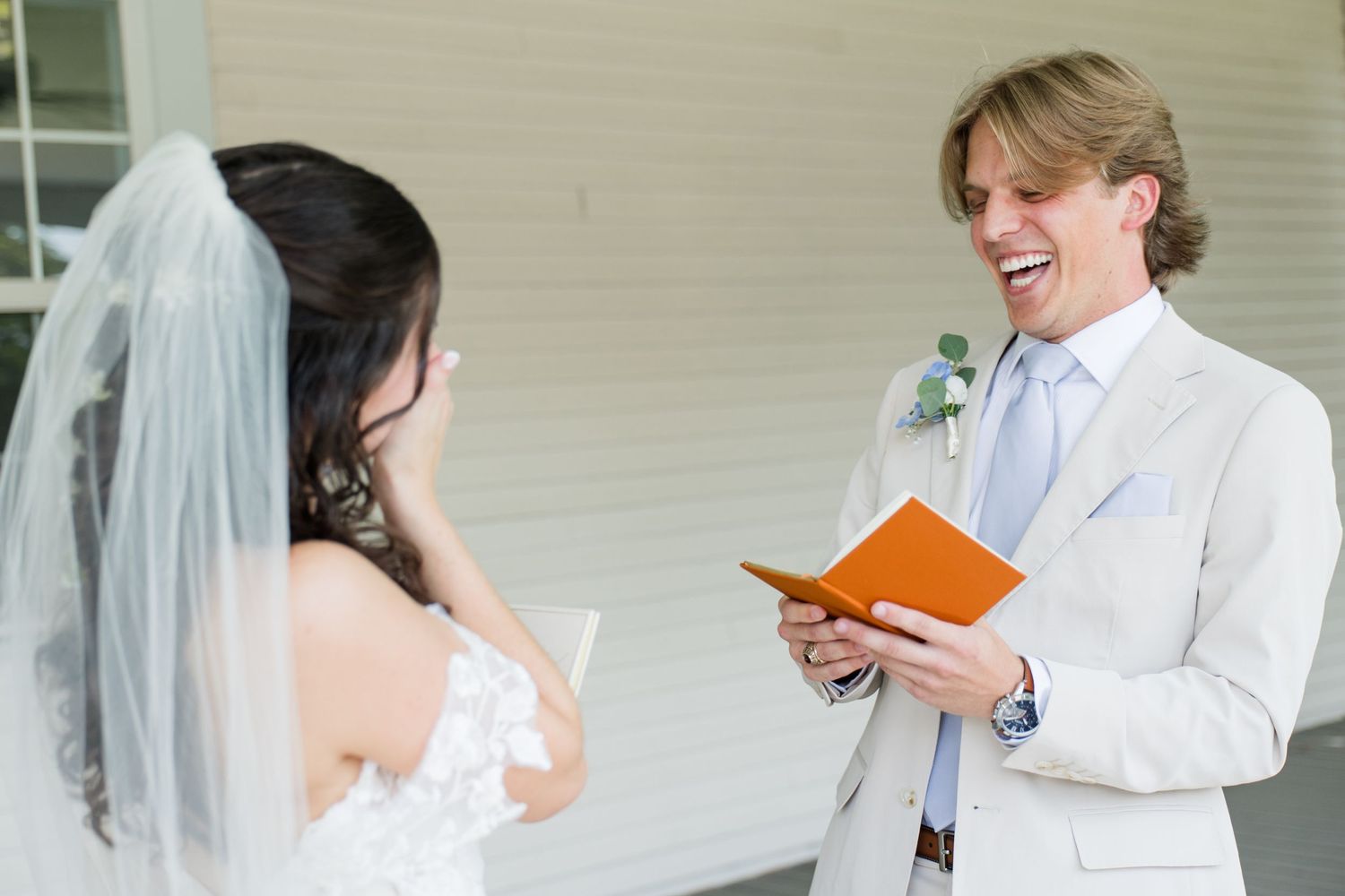 Groom in beige suit reads vows from orange book, smiling at bride in white lace dress and veil during wedding ceremony.