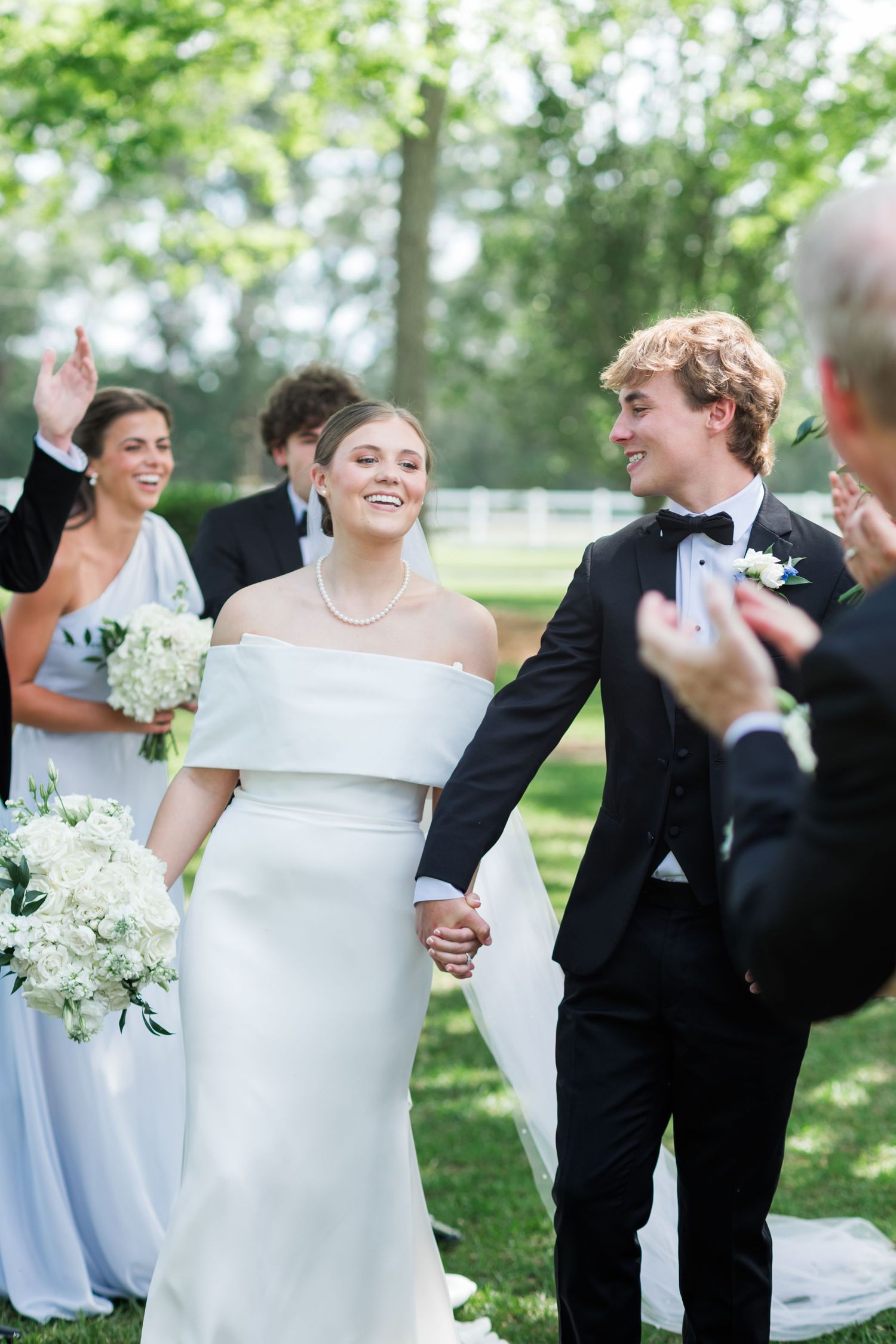 Joyful newlyweds walk down the aisle hand in hand, bride in off-shoulder gown holding white bouquet.