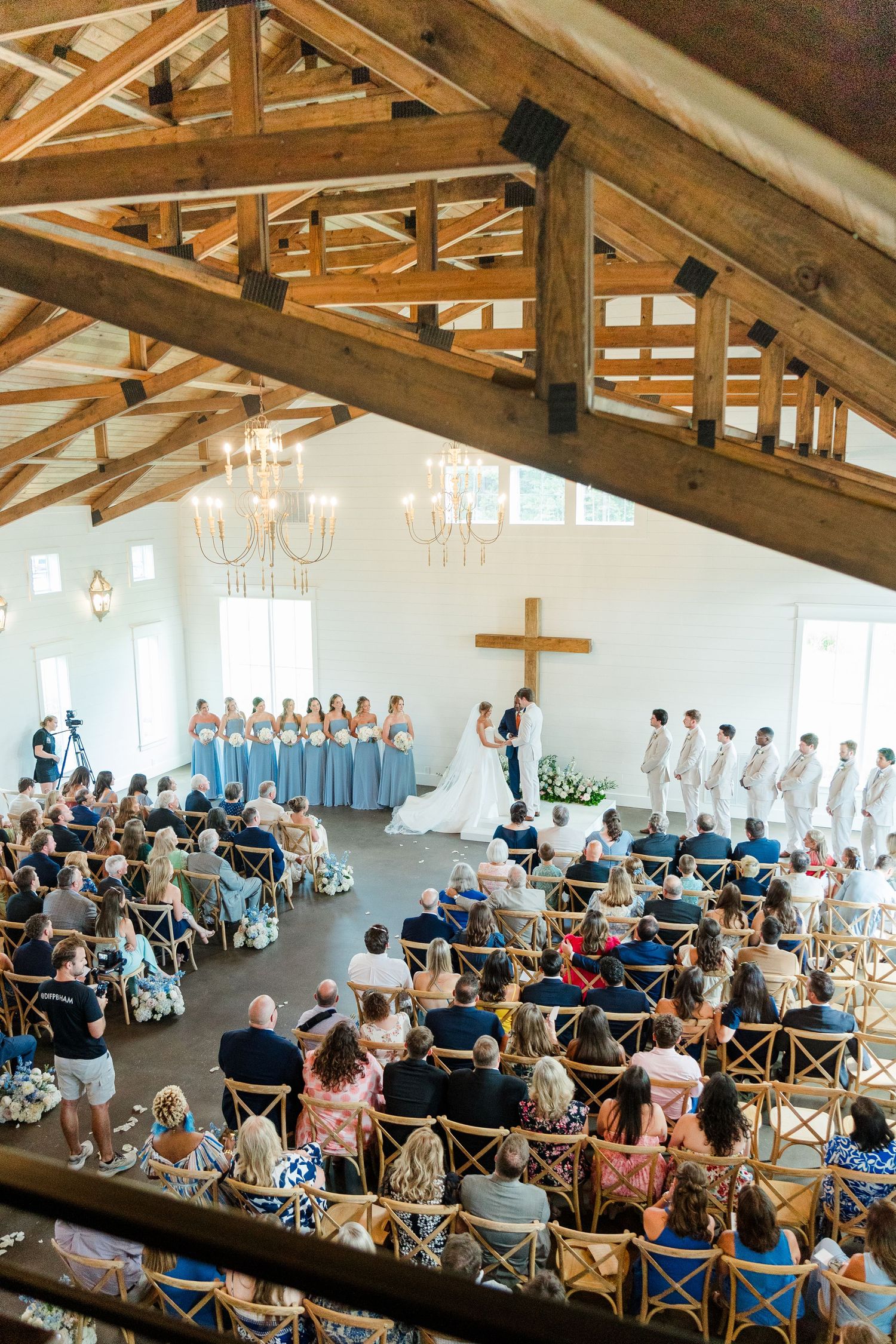 Elegant wedding ceremony in a white chapel with wooden beam ceiling, blue bridesmaid dresses, and a wooden cross at the altar.