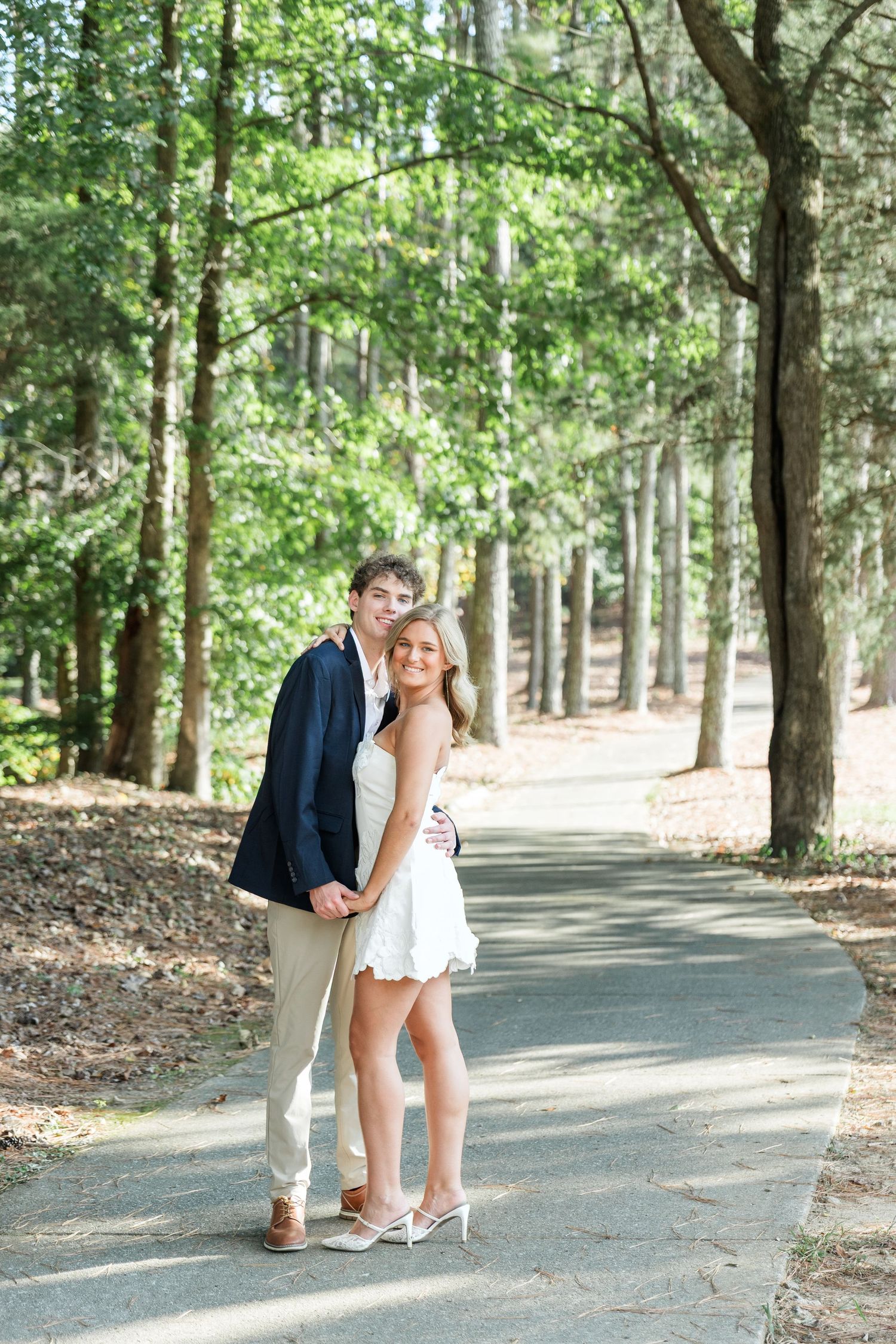 Couple holding hands on tree-lined path, woman in white dress, man in navy blazer.