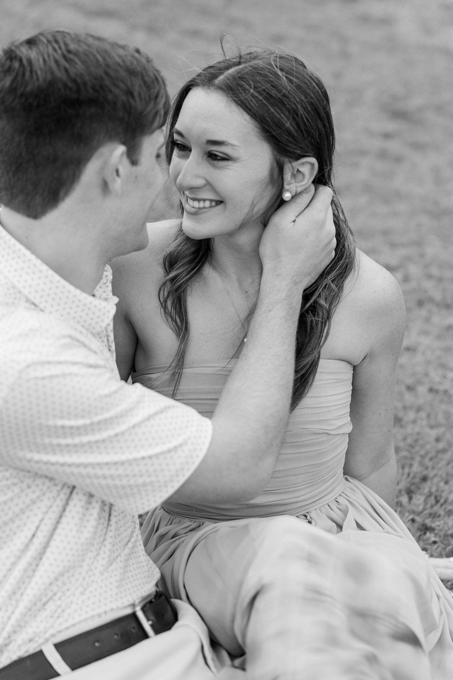 Black and white photo of a smiling woman looking at her partner as he gently touches her hair while seated on the grass.