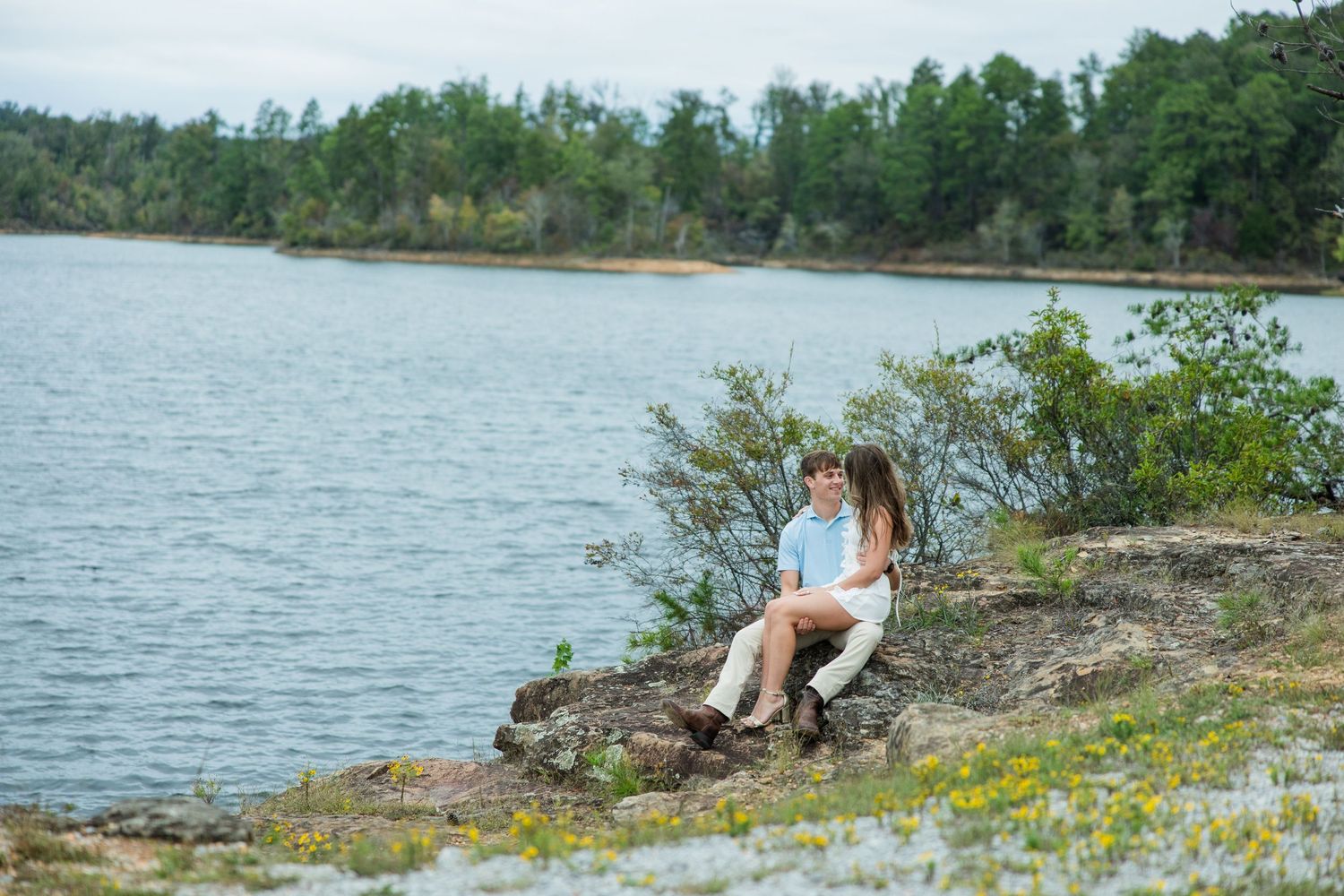 Couple sitting together on a rocky lakeside ledge with yellow wildflowers, overlooking calm water and green trees.