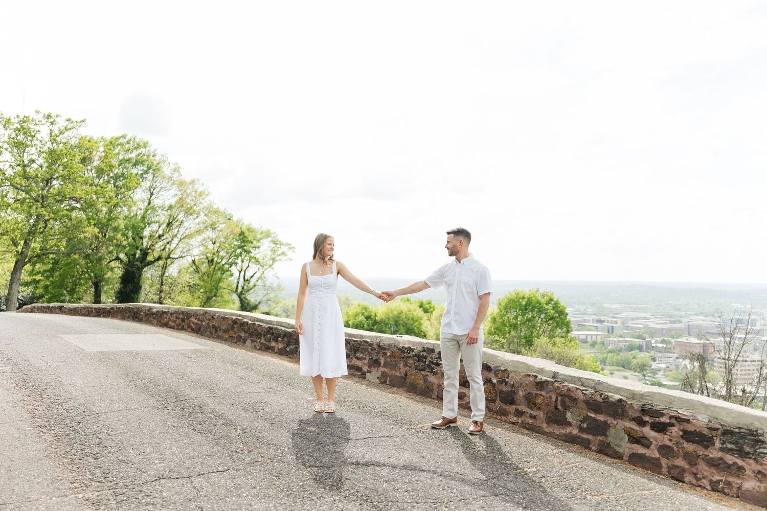 Couple holds hands on a scenic overlook road, woman in white dress, man in white shirt, with a valley view behind them.