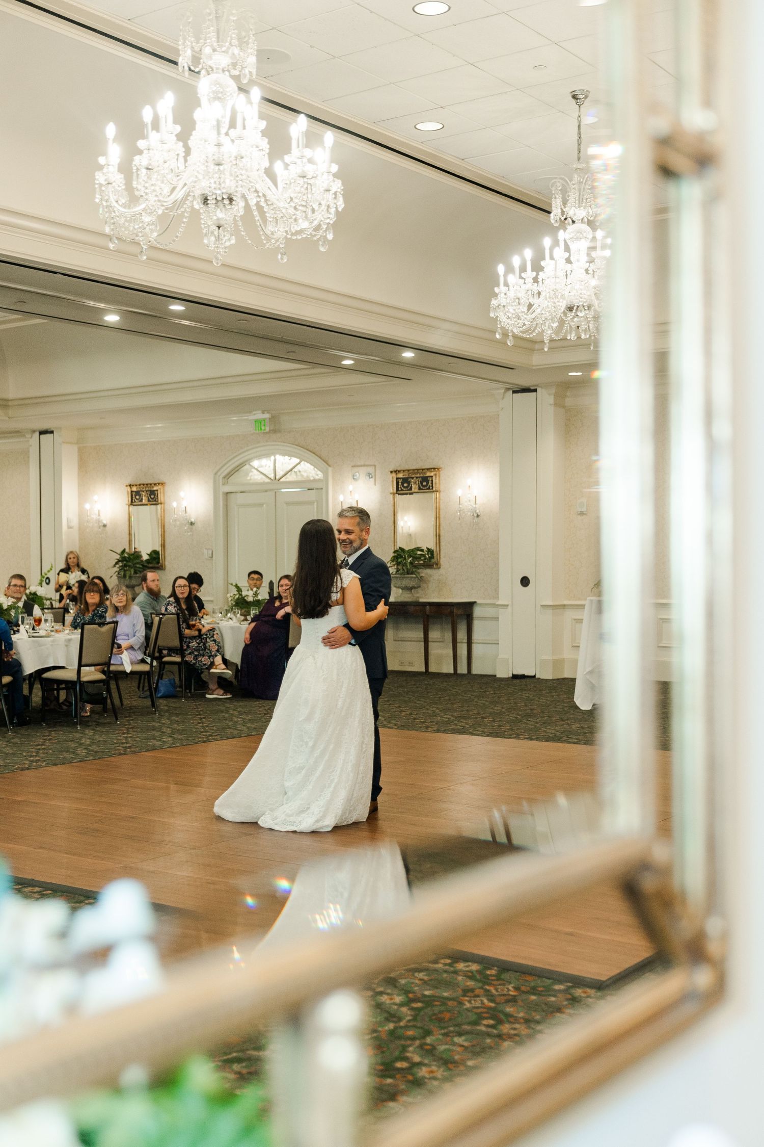 Bride and groom share first dance in grand ballroom with crystal chandeliers reflected in ornate gold mirror.