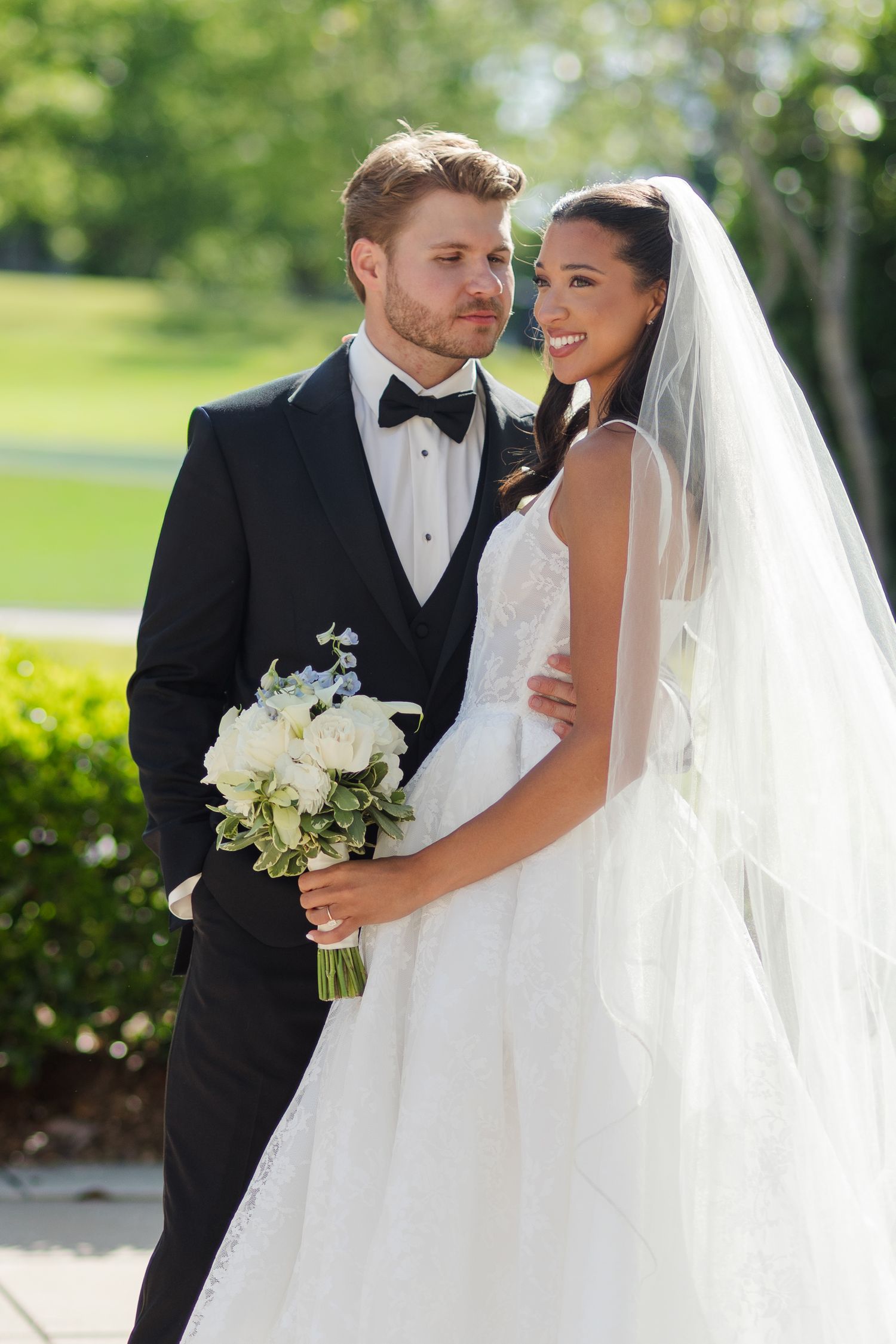 A bride and groom pose outdoors on their wedding day, she holds a white bouquet while wearing a veil and he wears a black tuxedo.