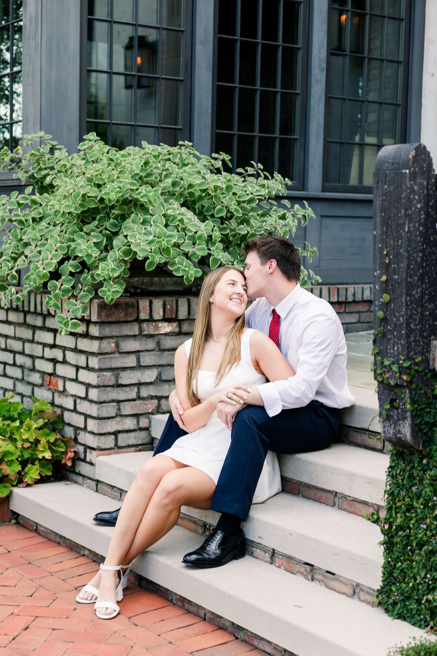 Couple sitting on brick steps, man in white shirt and red tie kissing woman in white dress who smiles joyfully.