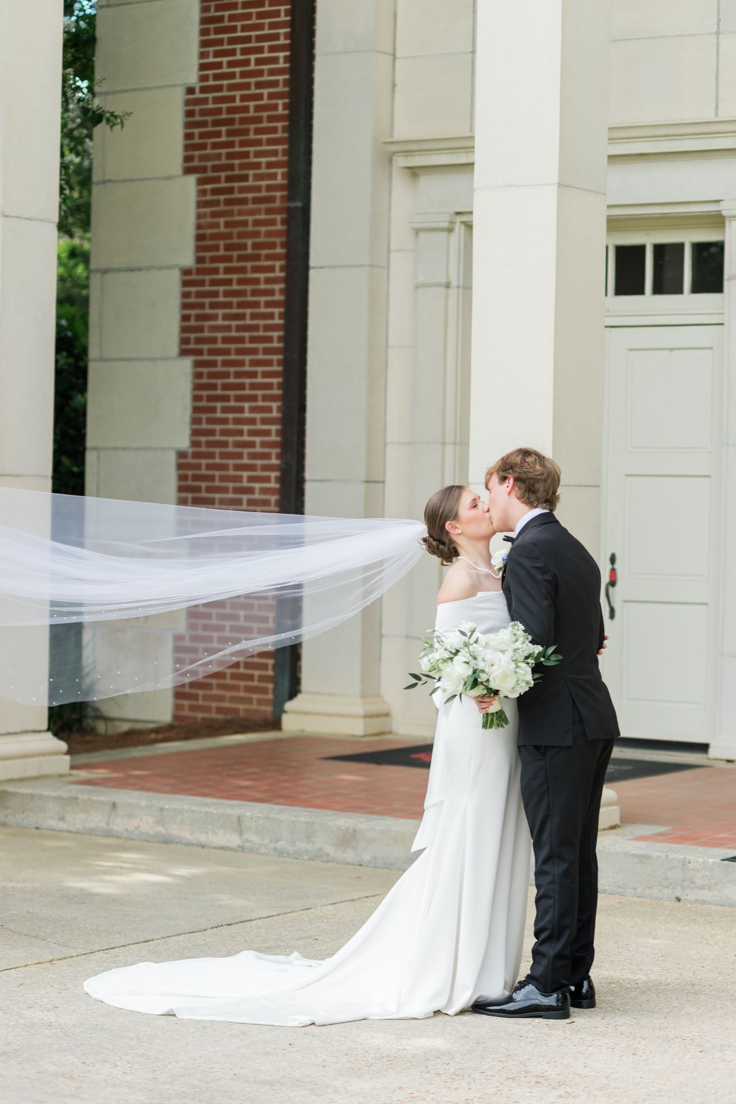 Bride and groom share a kiss outside elegant building as her long veil flows dramatically in the breeze.