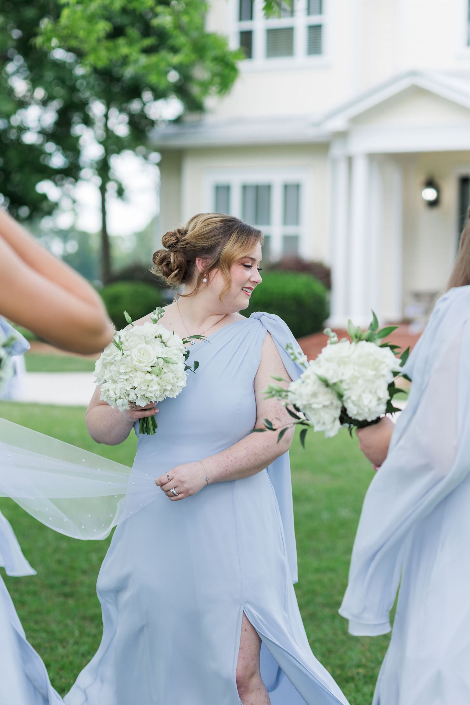 Smiling bridesmaid in light blue gown holding white hydrangea bouquet outdoors at a wedding celebration.