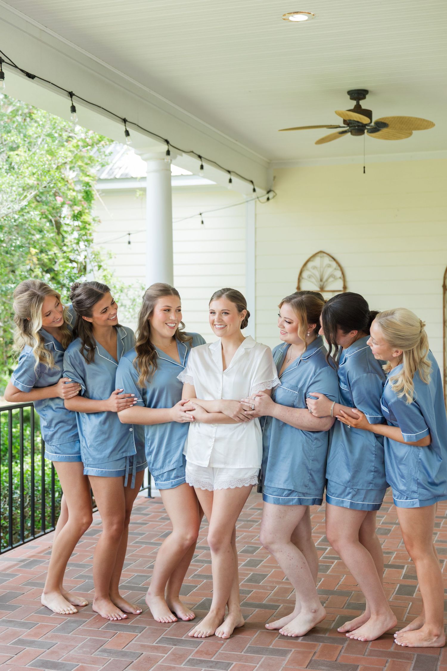 Bride in white pajamas surrounded by bridesmaids in blue on a porch before wedding ceremony.