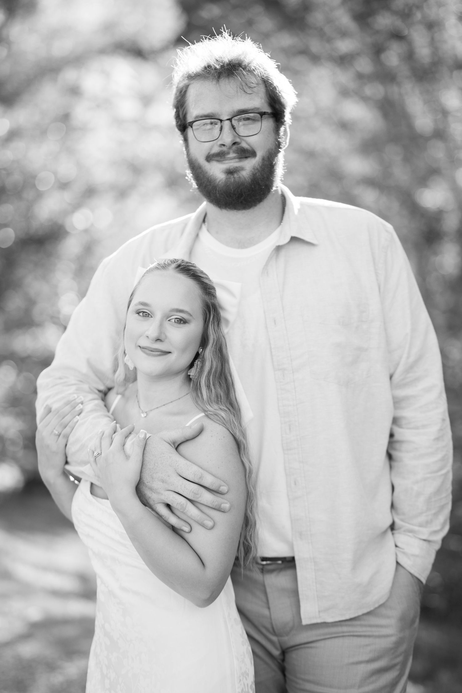Black and white portrait of a smiling couple embracing outdoors, man with beard and glasses hugging woman in white dress.