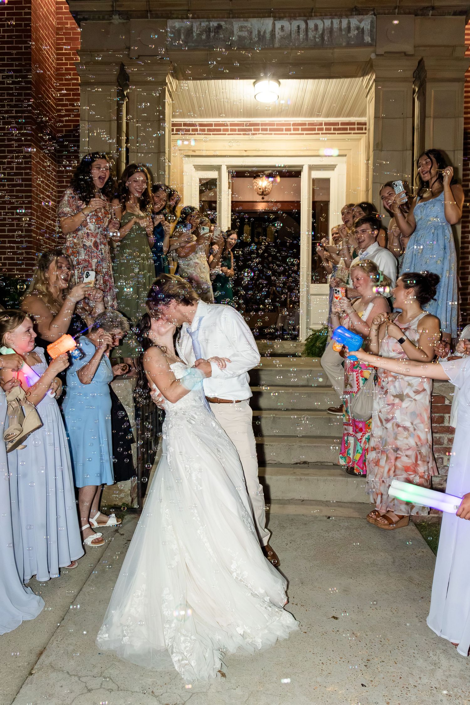 Bride and groom share romantic kiss during festive bubble exit outside The Emporium reception venue at night.