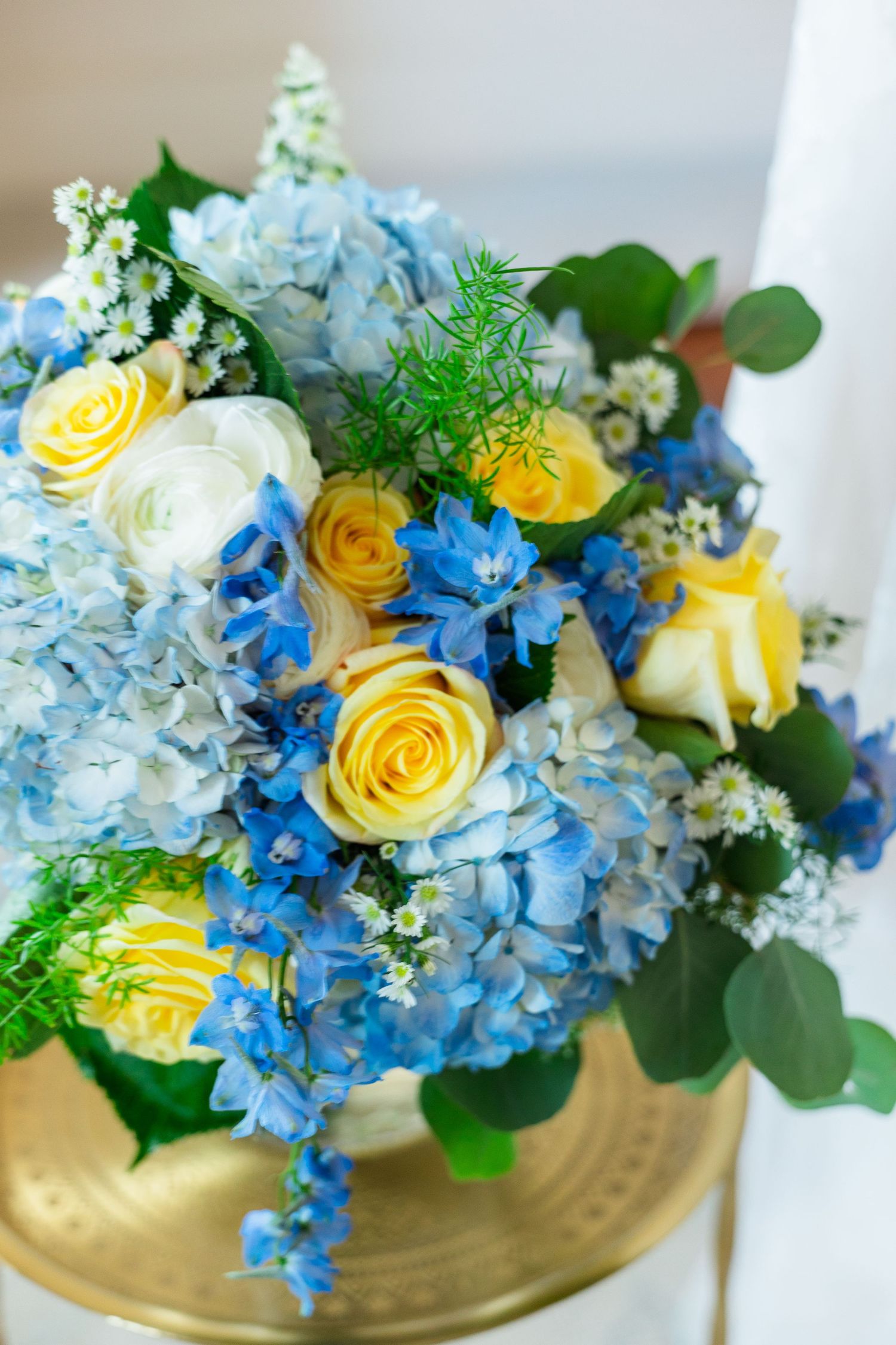 Blue hydrangea and yellow rose wedding bouquet arrangement in a gold vase with white flowers and greenery.