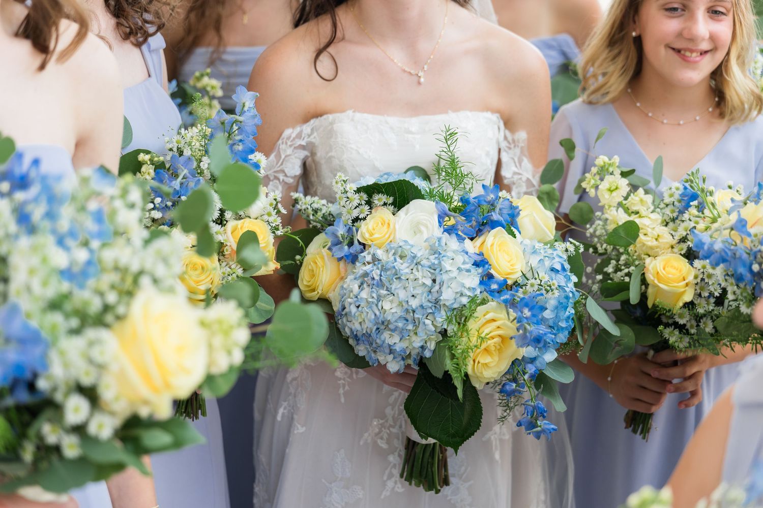 Bride and bridesmaids in blue dresses holding bouquets of yellow roses and blue hydrangeas at a wedding.