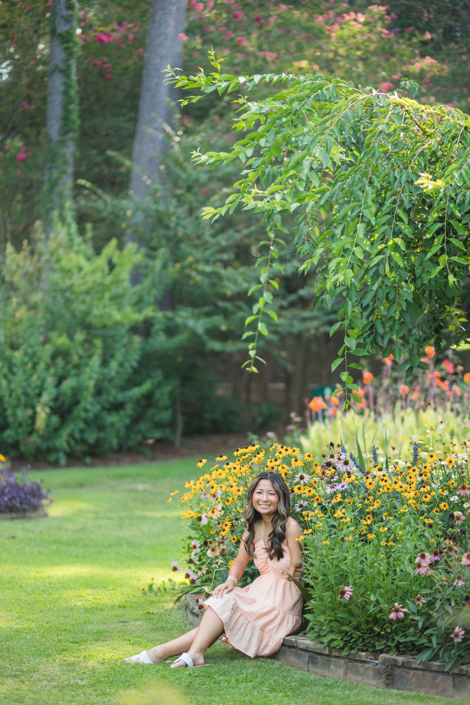 A smiling young woman in a peach dress sits on grass beside a colorful garden flower border.