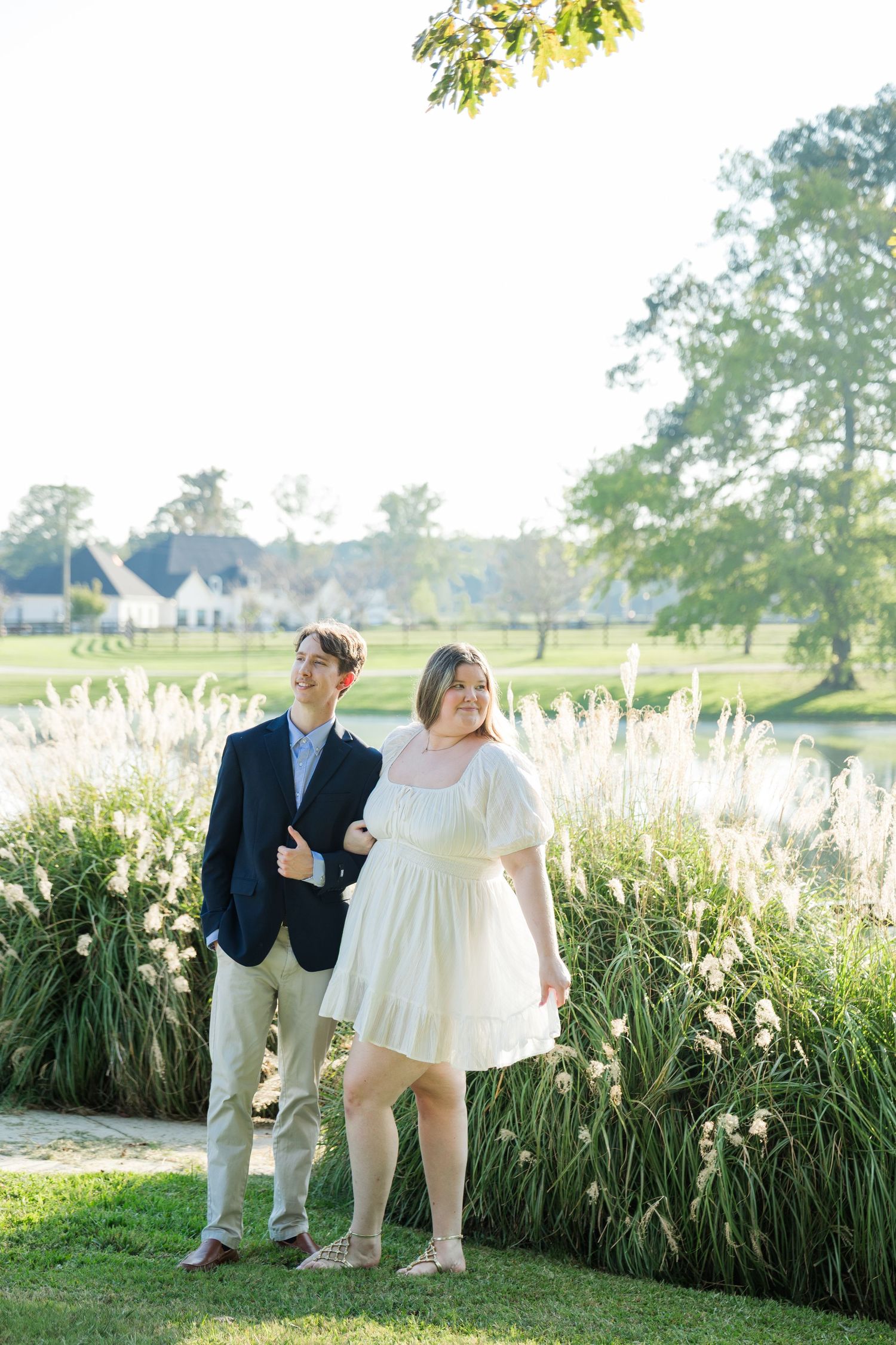 Couple posing outdoors near tall ornamental grasses with sunlit pond and estate in background.