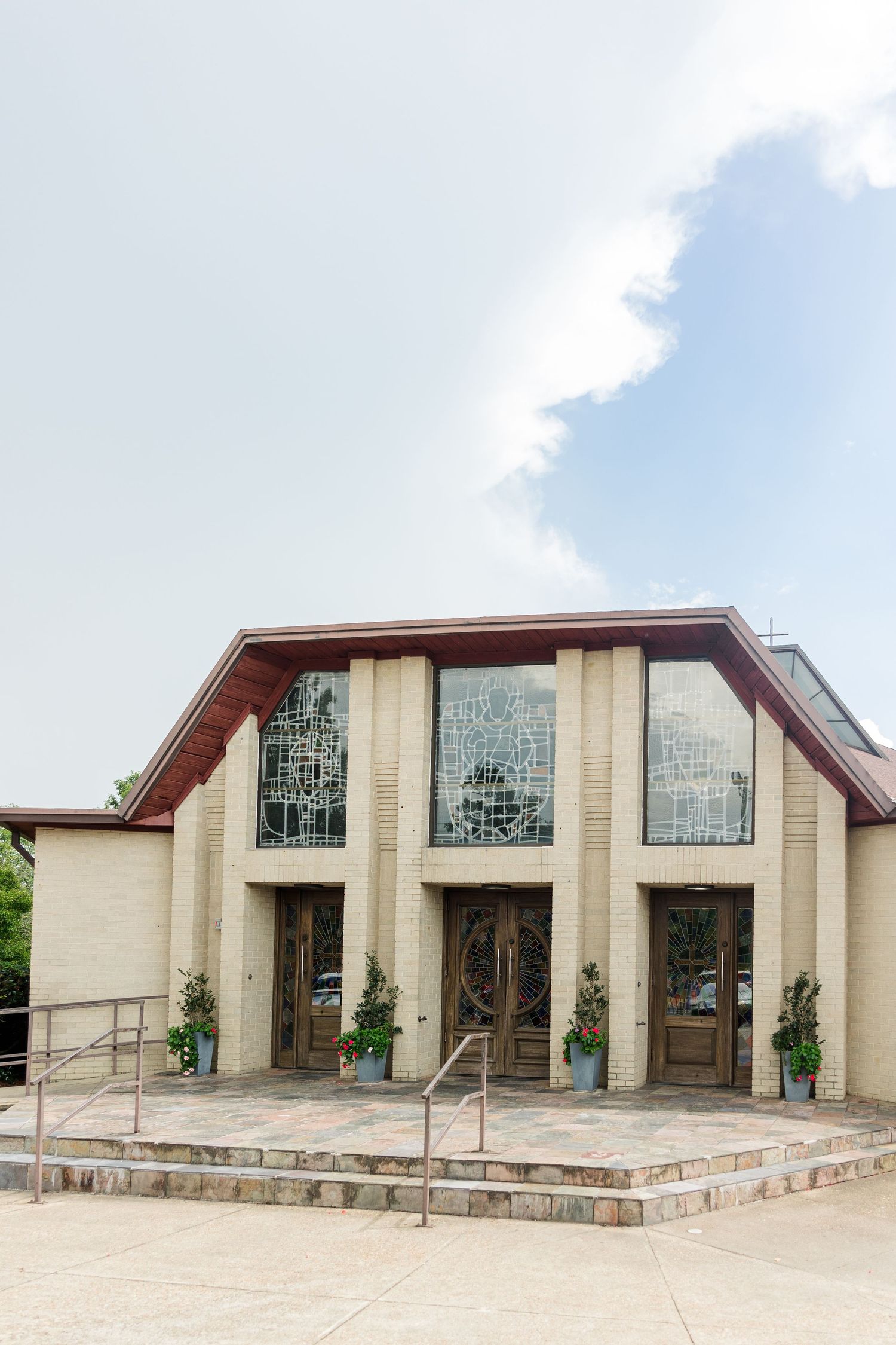 Modern church exterior with large stained glass windows, wooden doors, brick columns, and stone steps under a partly cloudy sky.