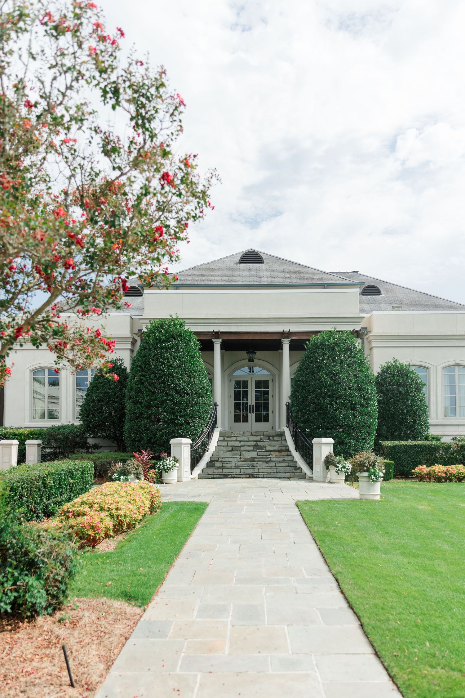 Elegant white Southern estate with manicured hedges, stone pathway, and blooming red flowers under cloudy skies.