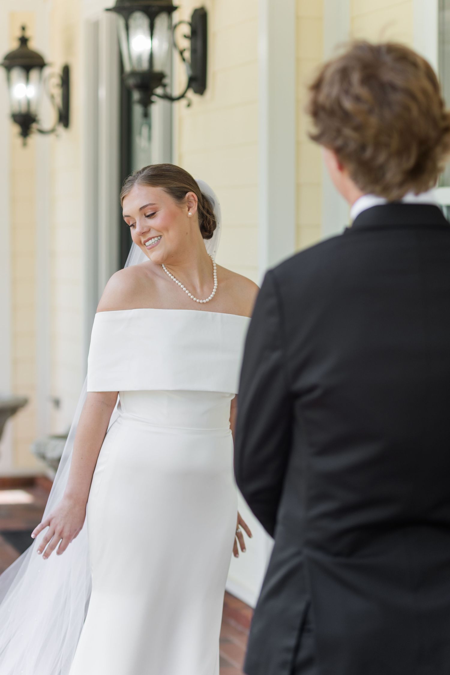 Smiling bride in off-shoulder white gown and pearl necklace faces groom in black tuxedo during wedding ceremony.