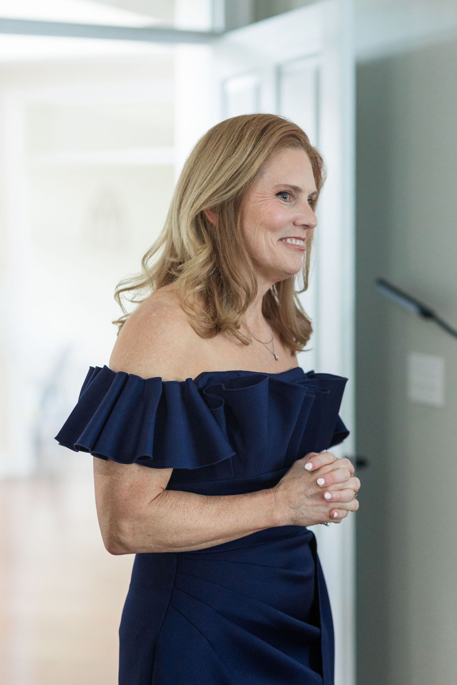 Elegant woman in navy off-shoulder ruffle dress smiling in a bright modern hallway.