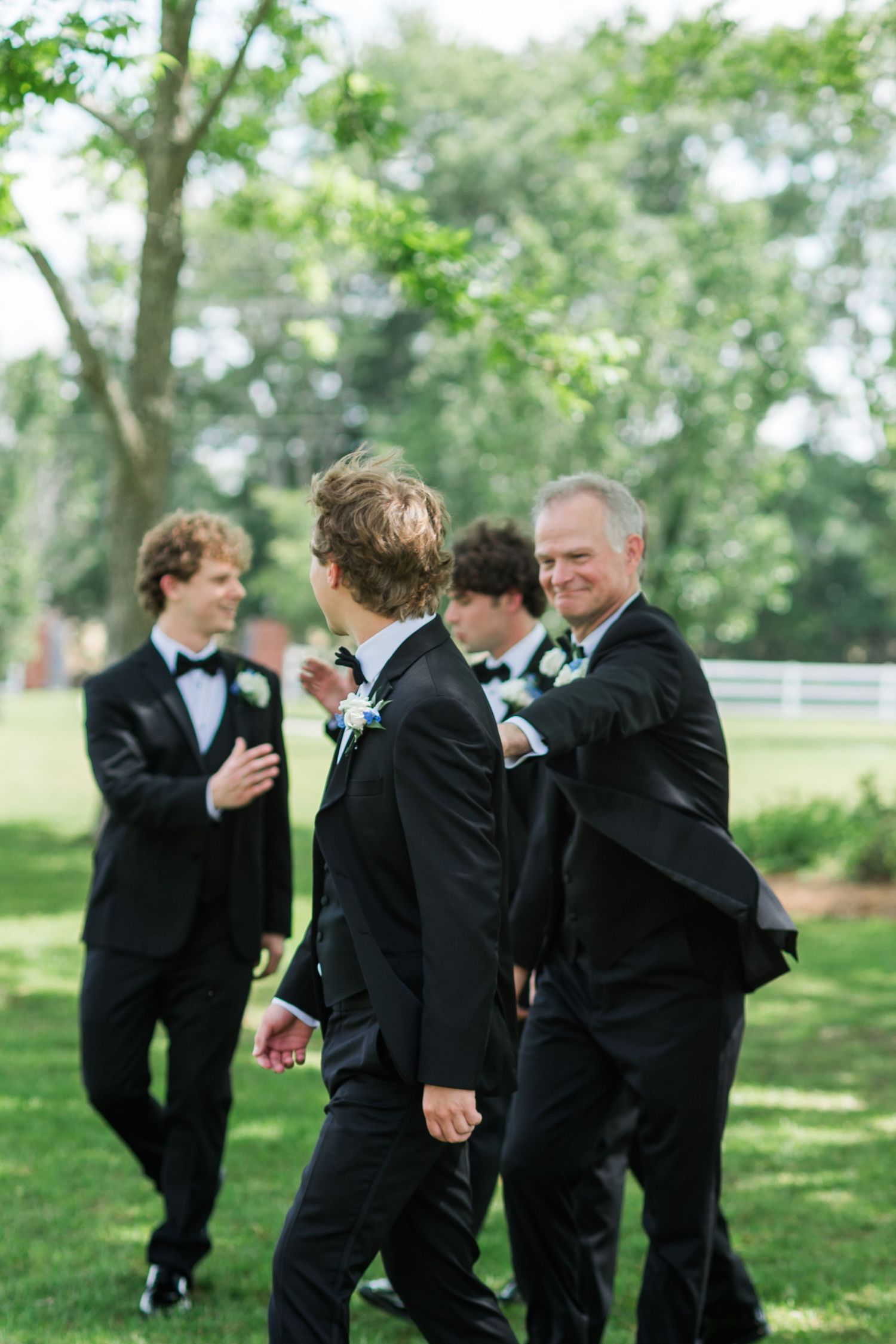 Groomsmen and older gentleman in black tuxedos walk and laugh together on green lawn at outdoor wedding.