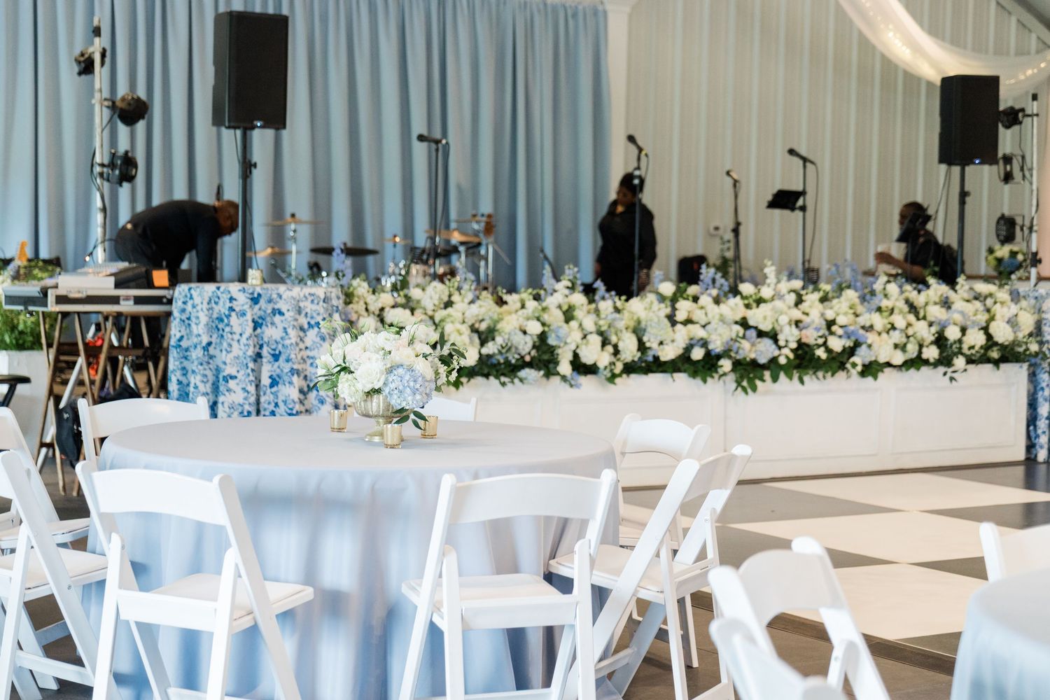 White wedding reception tent with round tables, white folding chairs, blue linens, and lush floral centerpieces on dance floor.