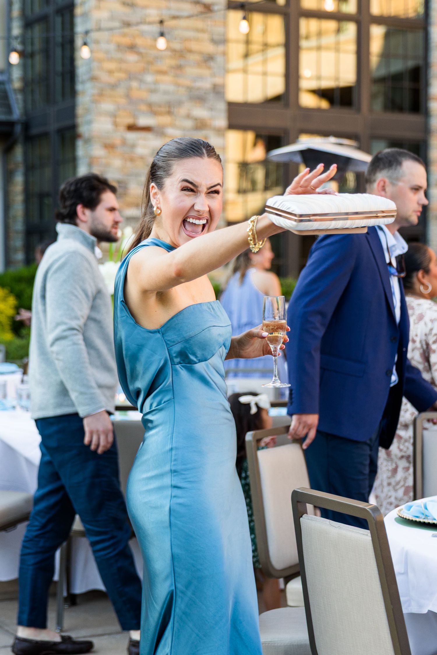 Woman in blue gown laughing at outdoor party, holding champagne glass at elegant evening social event.