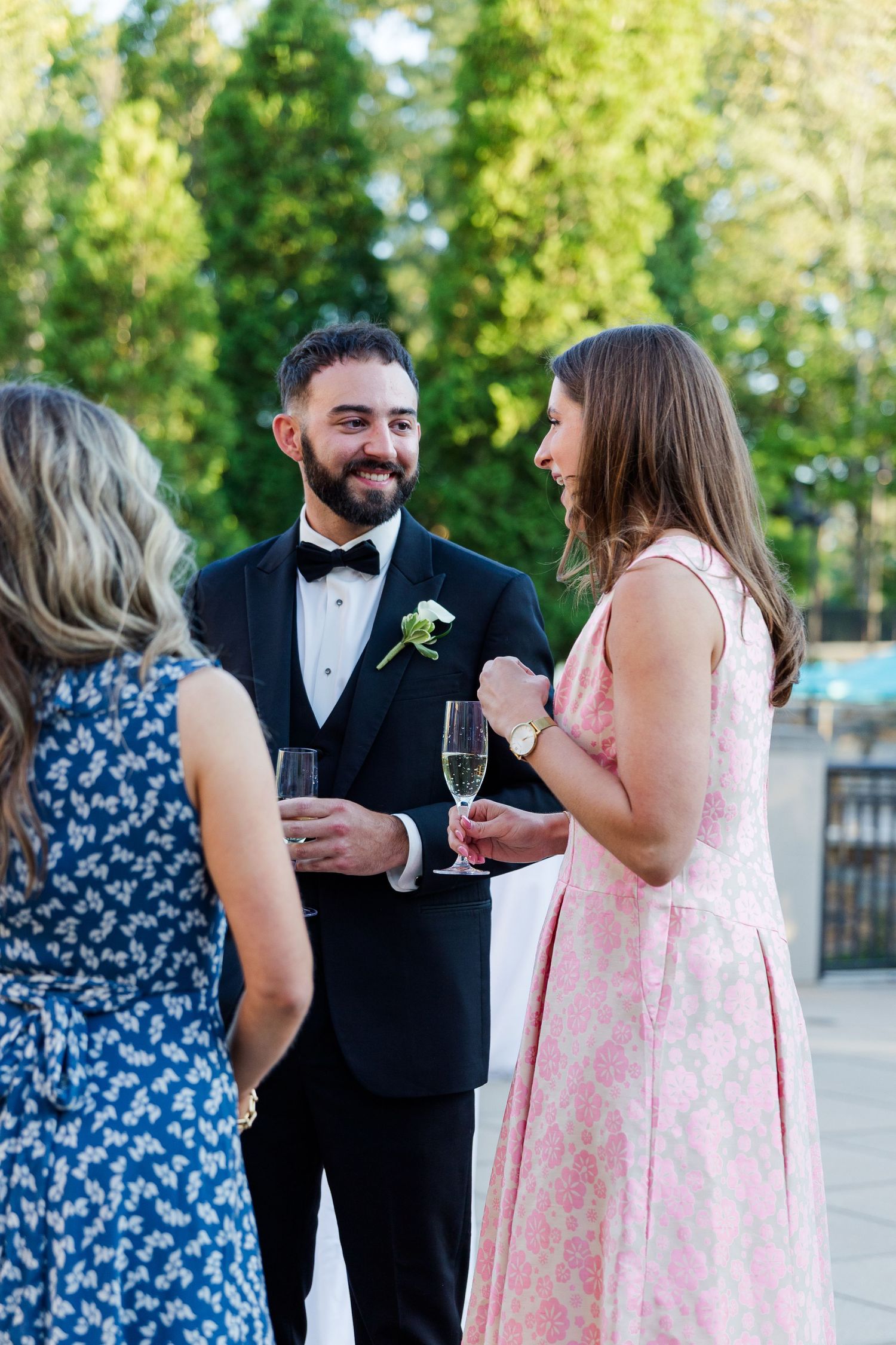 Groom in black tuxedo with boutonniere smiles while chatting with guests in elegant dresses at outdoor wedding reception.