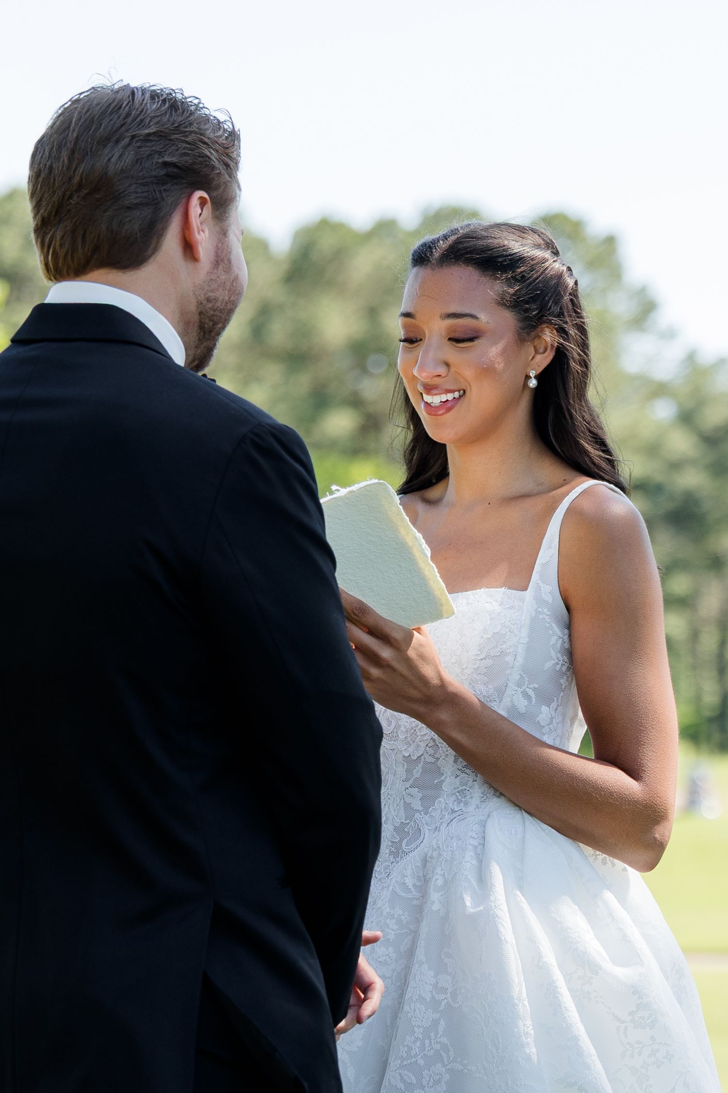 Smiling bride in white gown reads wedding vows from a small book while standing outdoors with groom in black suit.