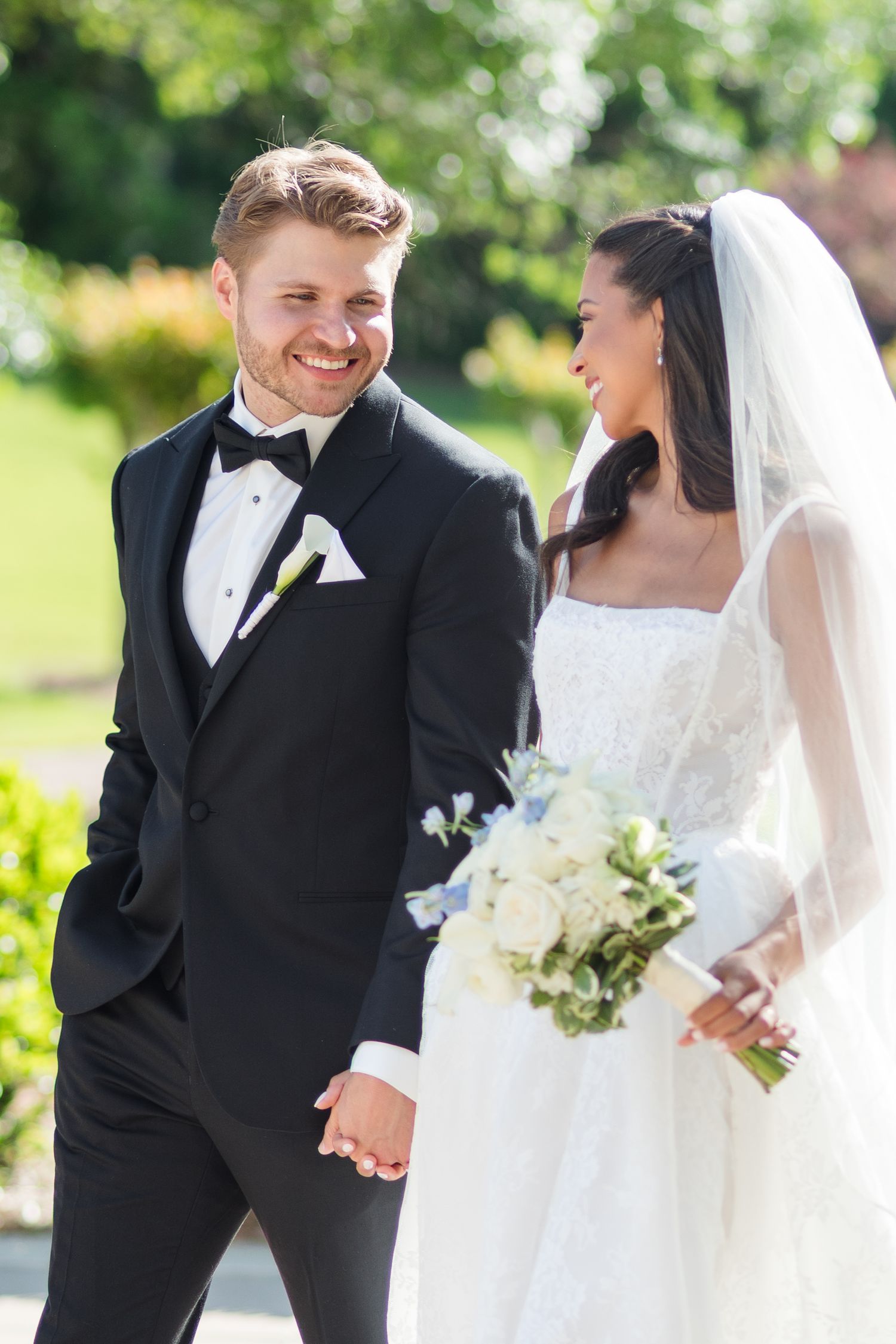 A smiling groom in a black tuxedo holds hands with his bride in a white gown and veil, walking outdoors on their wedding day.