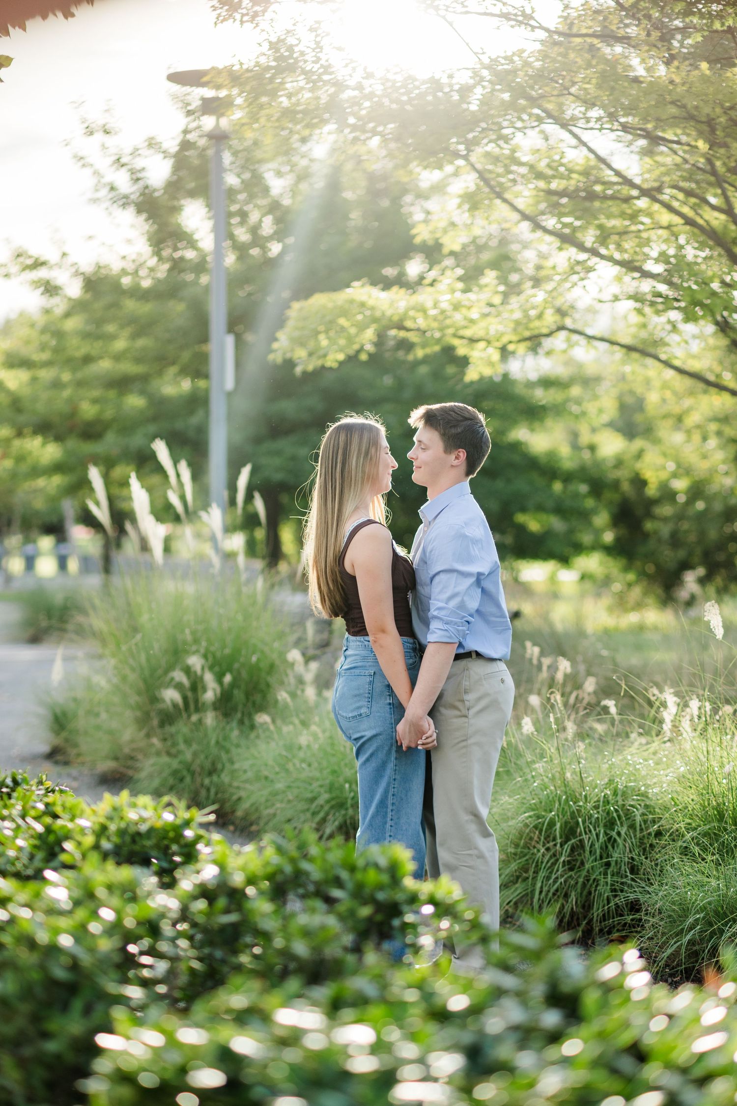 Couple holding hands facing each other in sunlit garden surrounded by ornamental grasses and greenery.