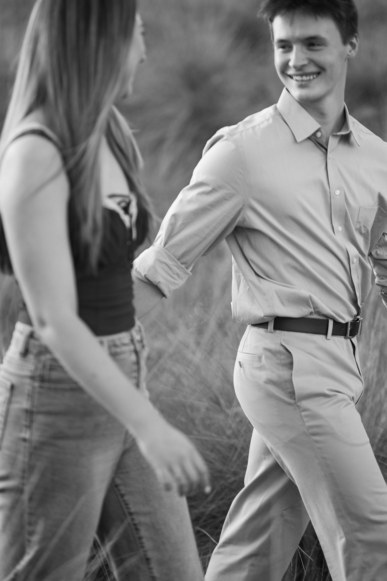 A black and white photo of a smiling young man walking with a woman through tall grasses outdoors.