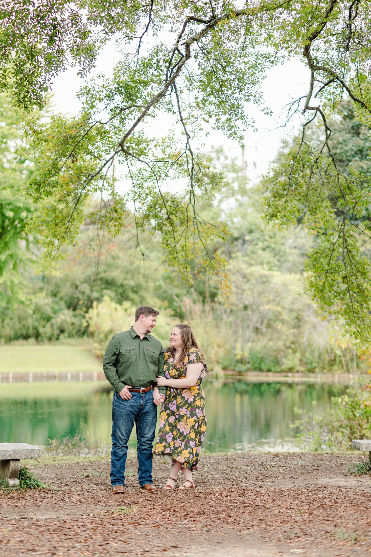Couple smiling at each other in floral and green outfits by a serene pond surrounded by lush trees.