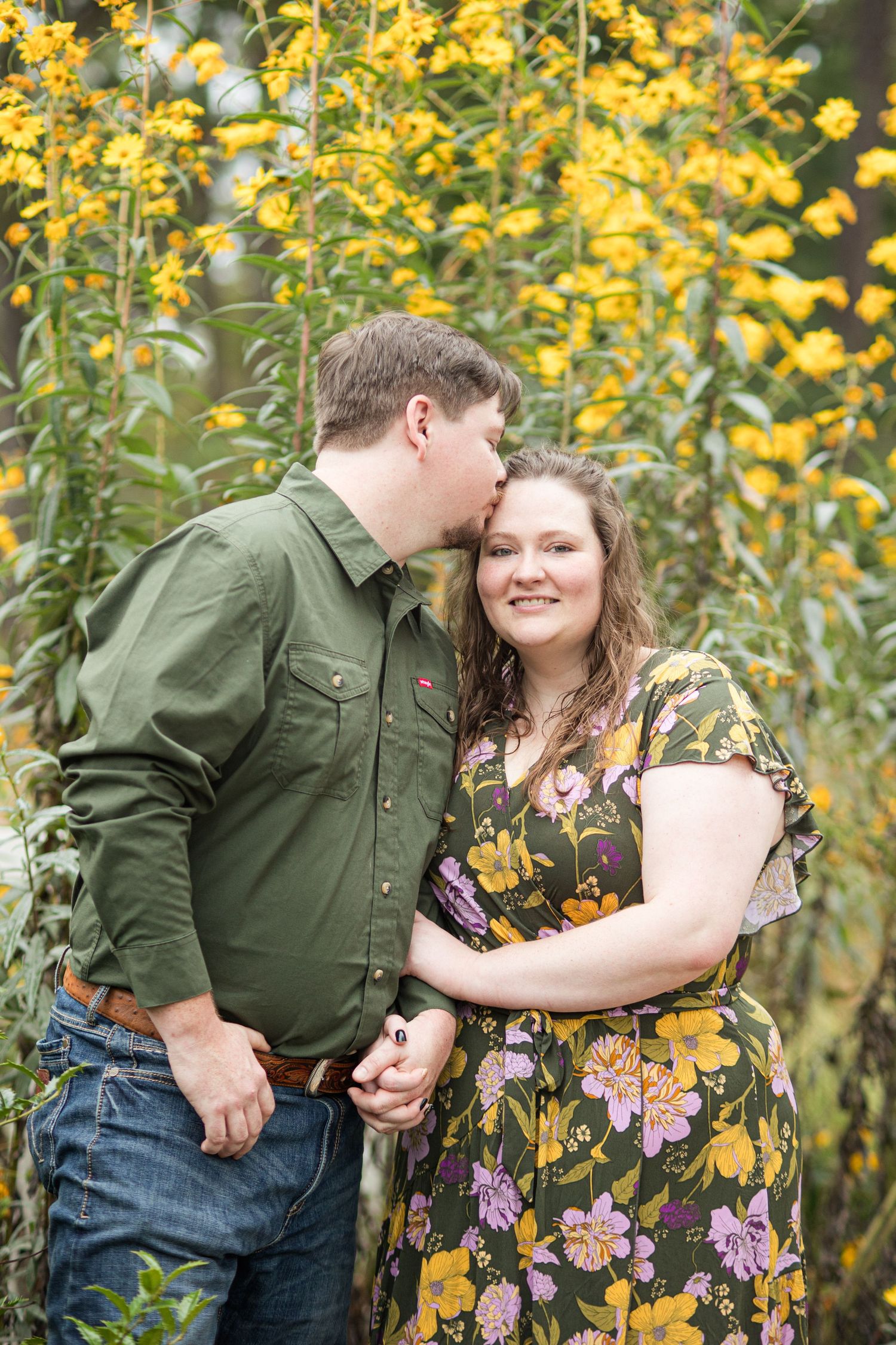 A couple shares a tender kiss among vibrant yellow wildflowers, the man in green shirt and jeans, woman in floral dress.