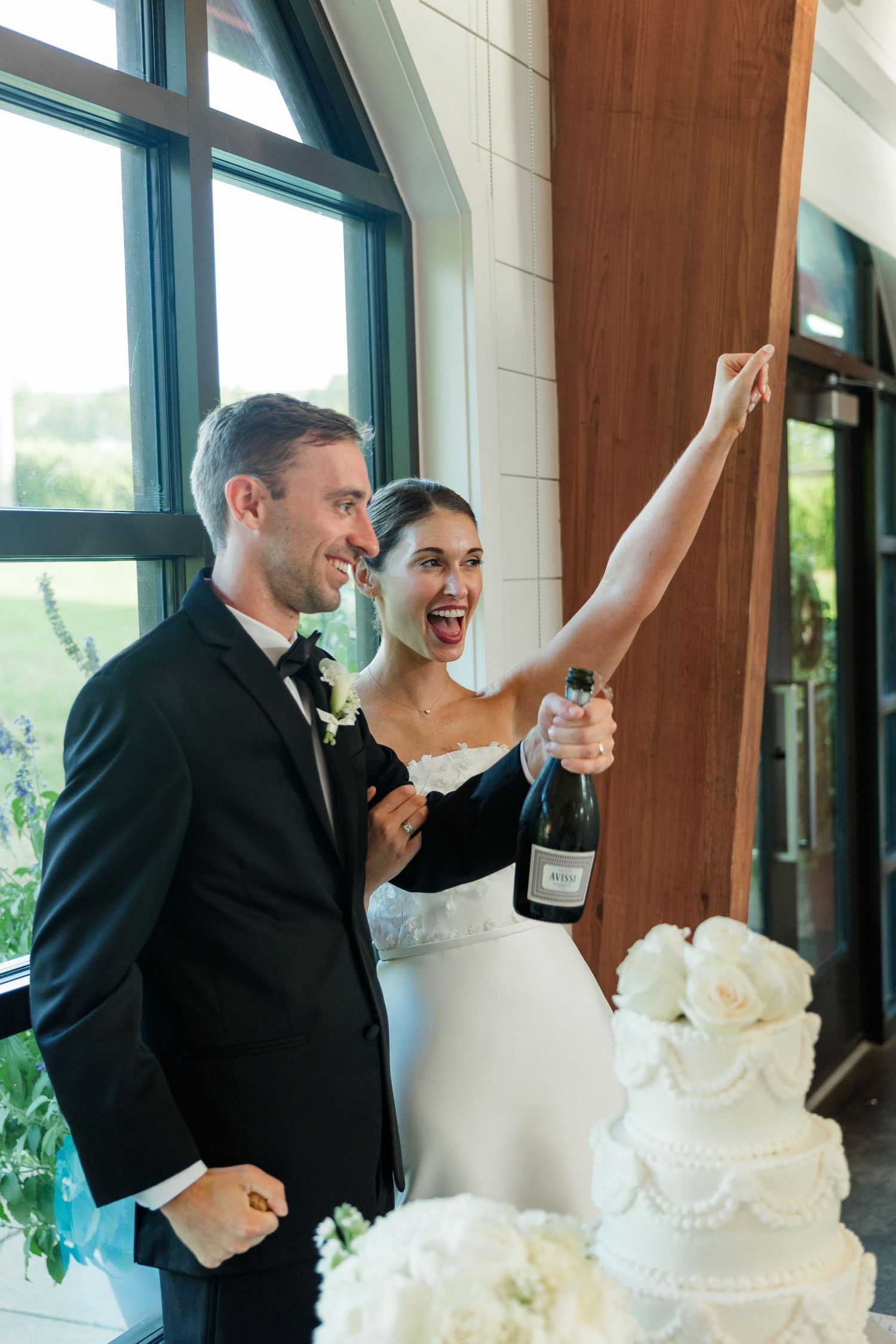 Bride holding champagne bottle and groom in tuxedo celebrate at wedding reception near white tiered cake.