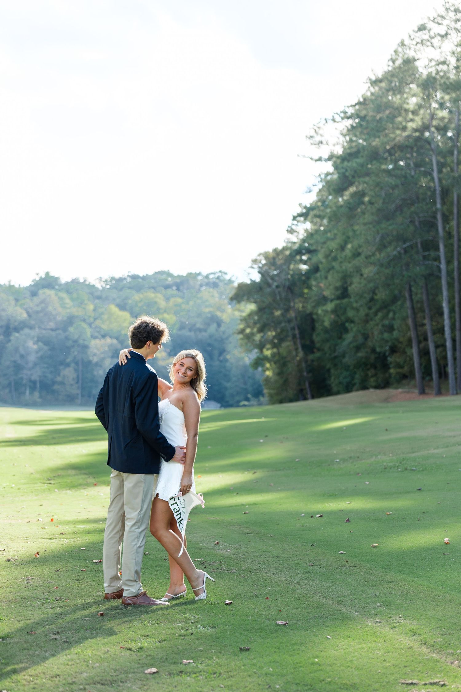 Couple embracing on a sunny golf course fairway surrounded by tall trees, woman holding a pennant flag.