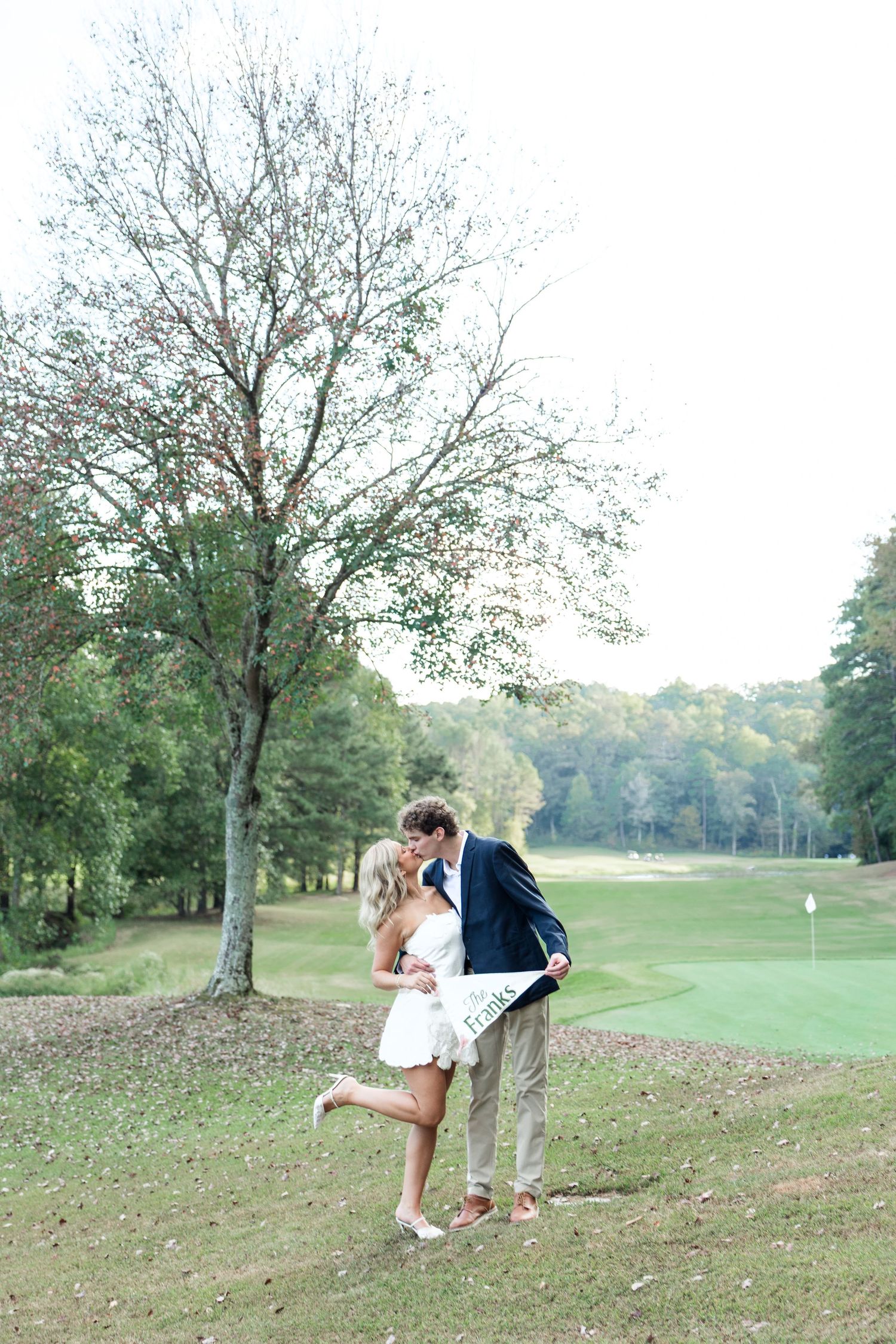 Couple sharing a kiss on a golf course at dusk, bride lifting her leg while holding a white engagement pennant.