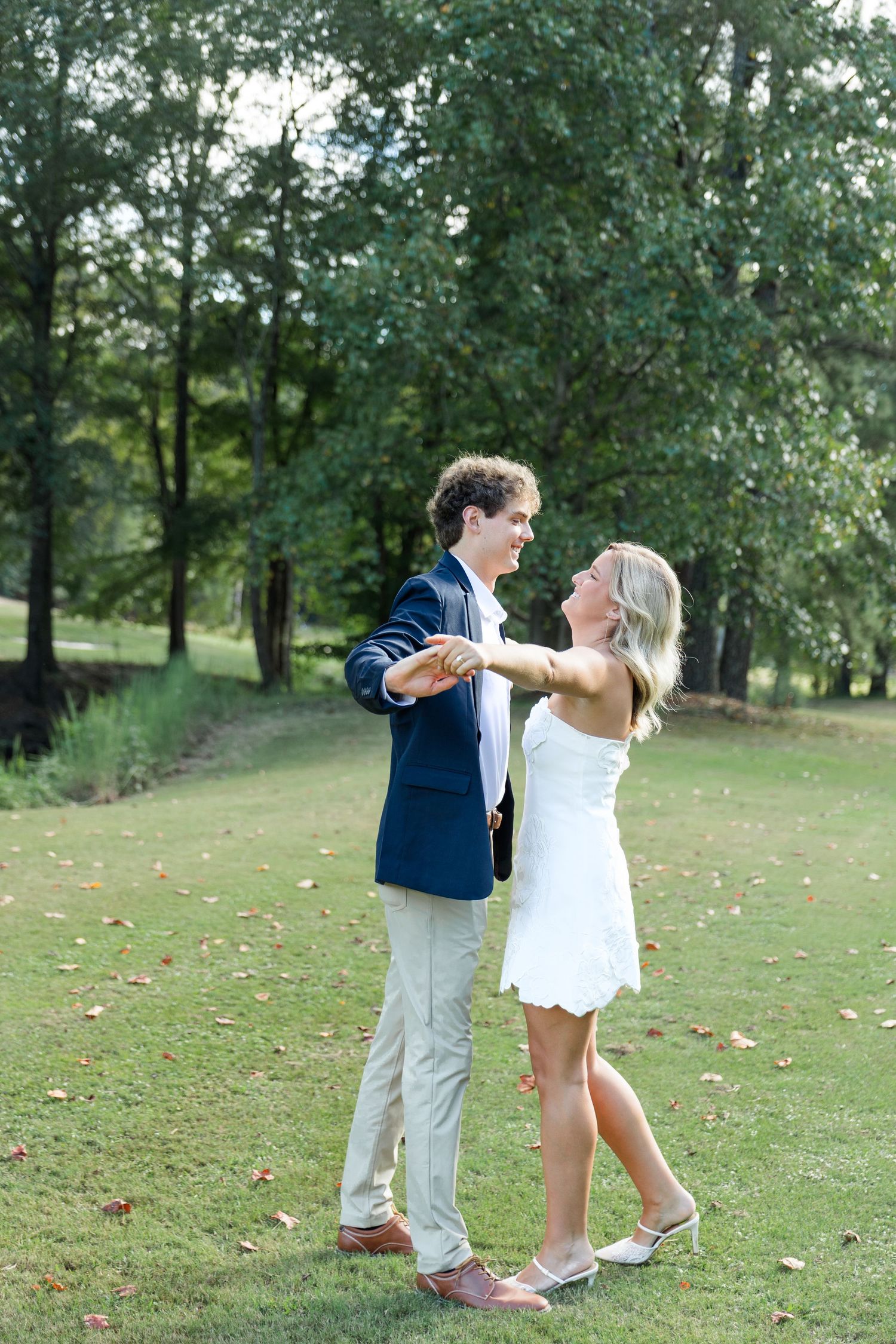 Young couple dancing and holding hands on a green lawn with lush trees in the background.