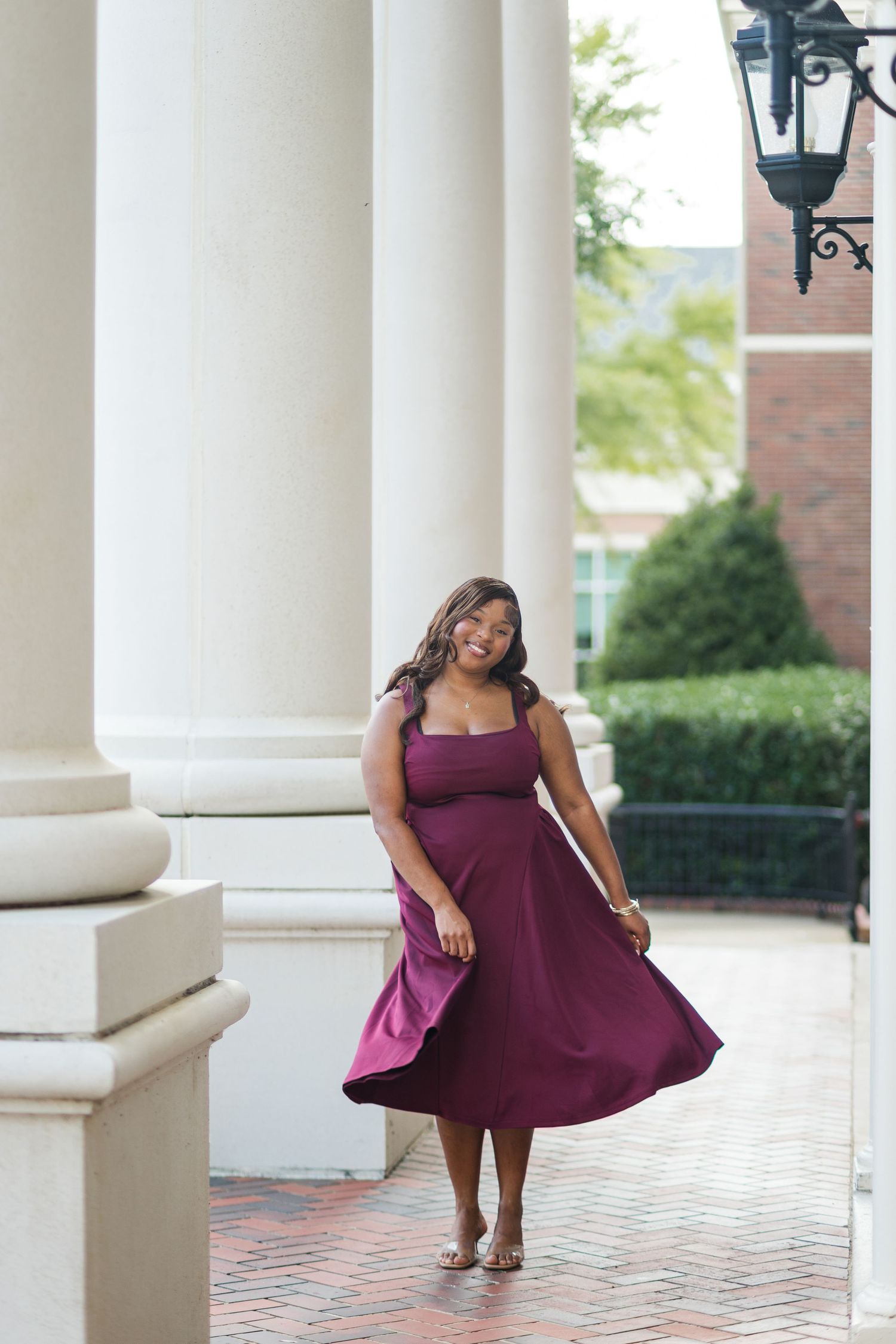 Smiling graduate in flowing burgundy dress posing between white columns on university campus.