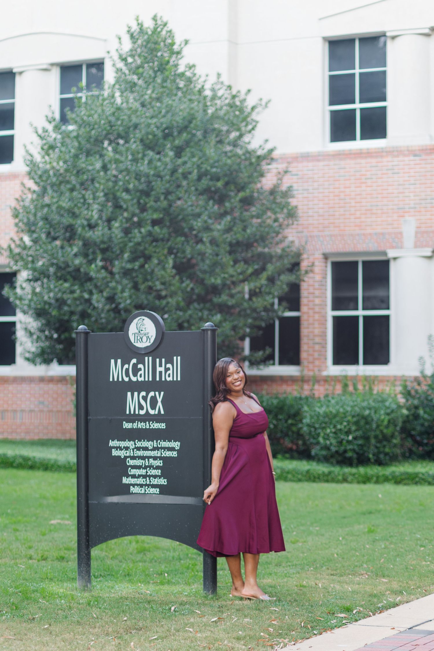 Smiling graduate in burgundy dress poses beside McCall Hall MSCX sign on college campus.