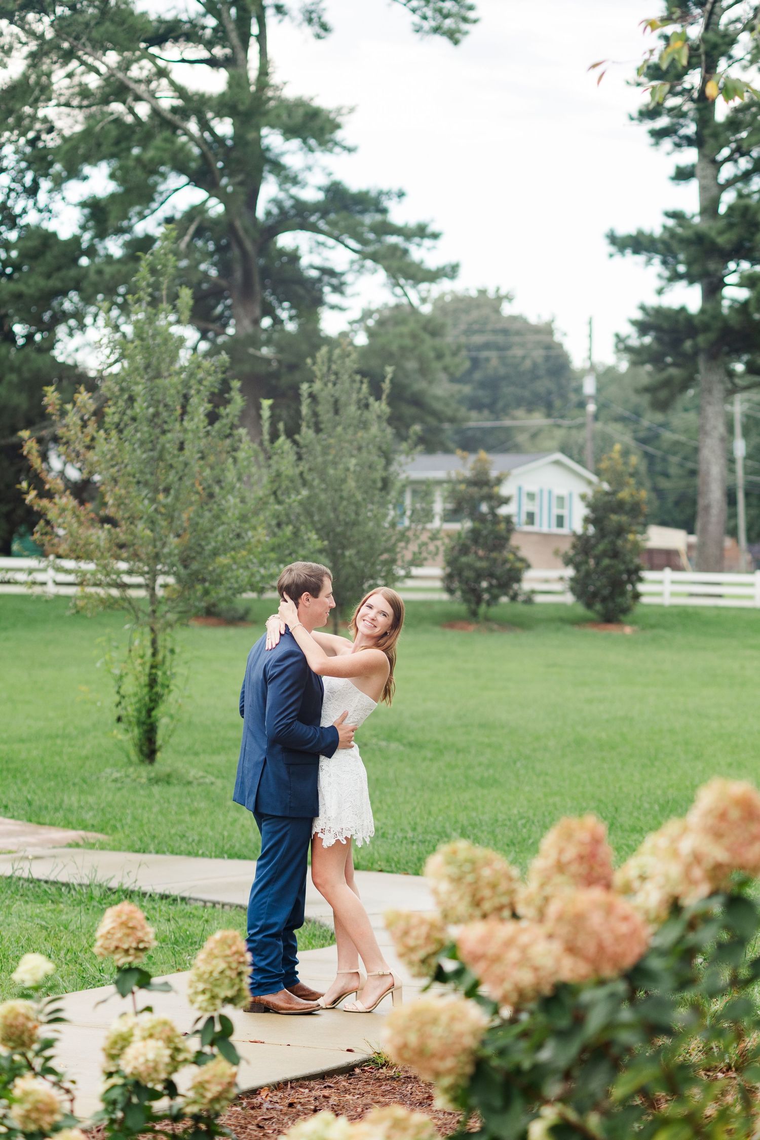 Couple embracing in a lush garden with hydrangeas and a white fence at a charming estate.