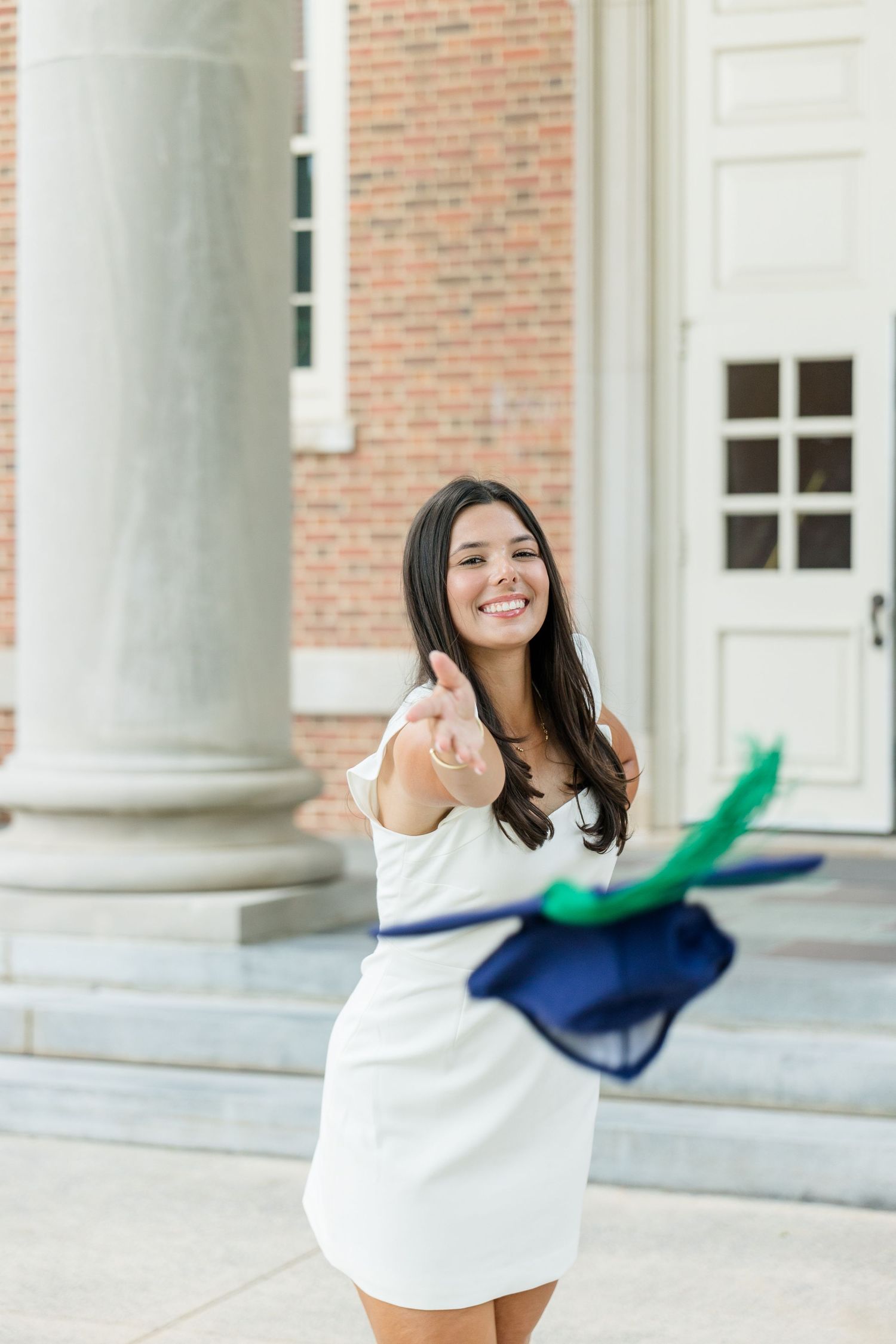 Happy female graduate in white dress tossing navy and green graduation cap in front of brick building.
