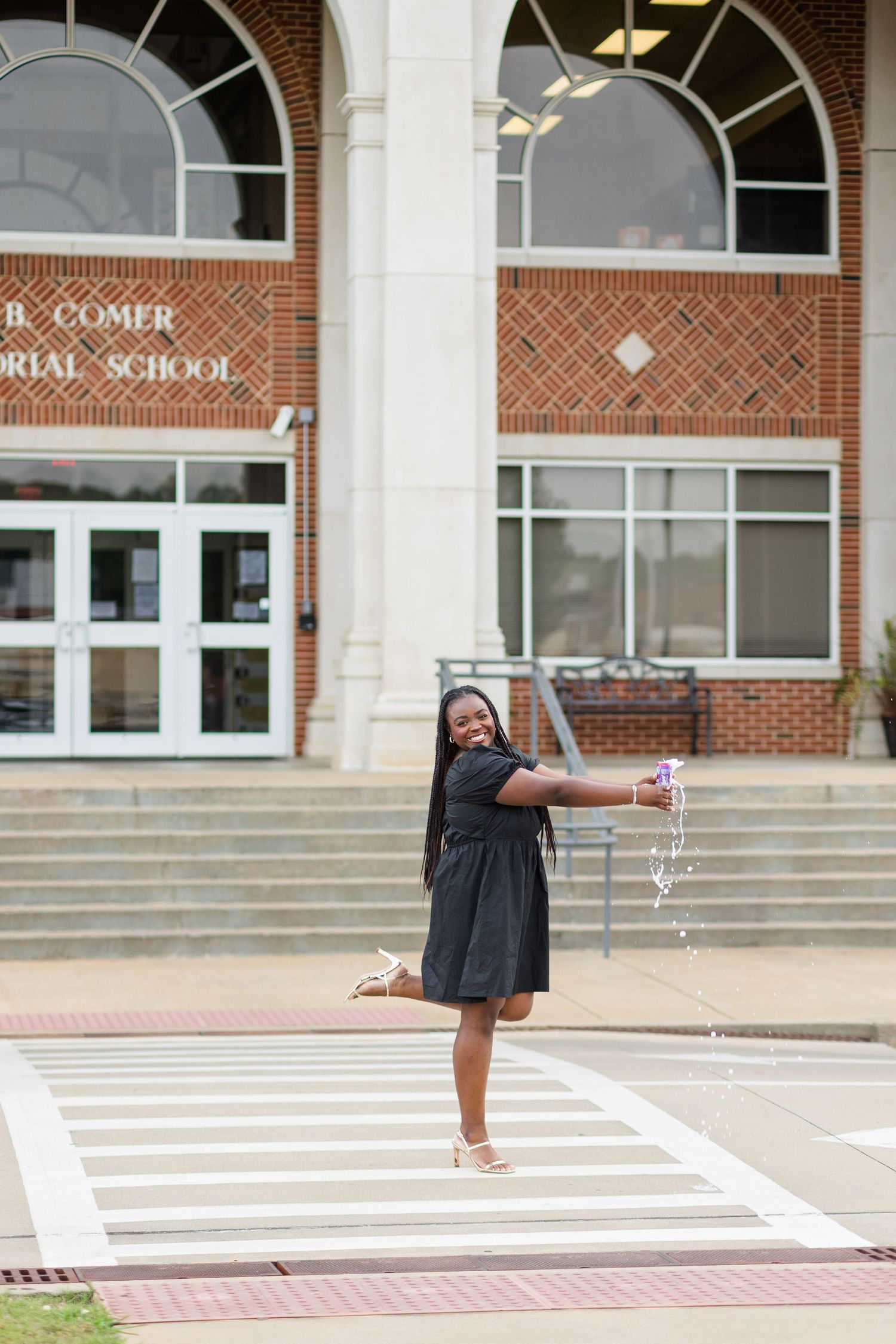 Joyful young Black woman in black dress dancing on crosswalk in front of brick school building.