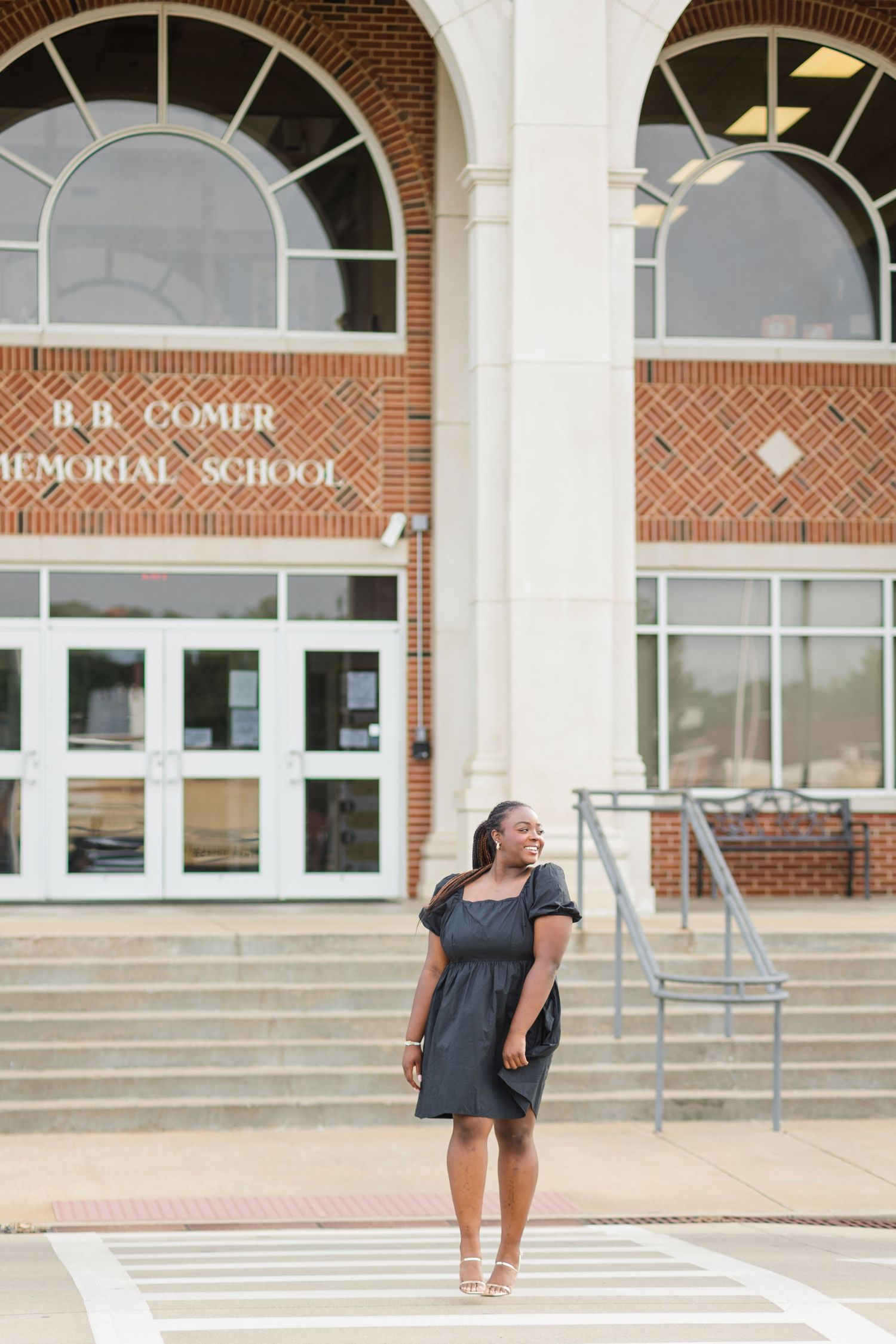 A young Black woman in a dark dress poses confidently in front of B.B. Comer Memorial School entrance.