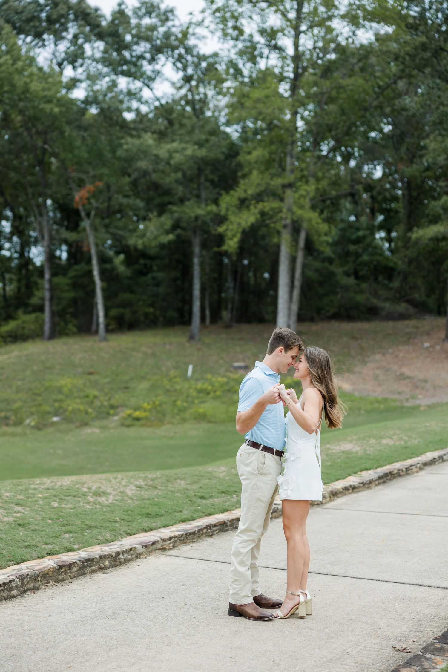 Couple in casual attire sharing a close dance on a stone path beside a green golf course lawn and tall trees during an engagement session.