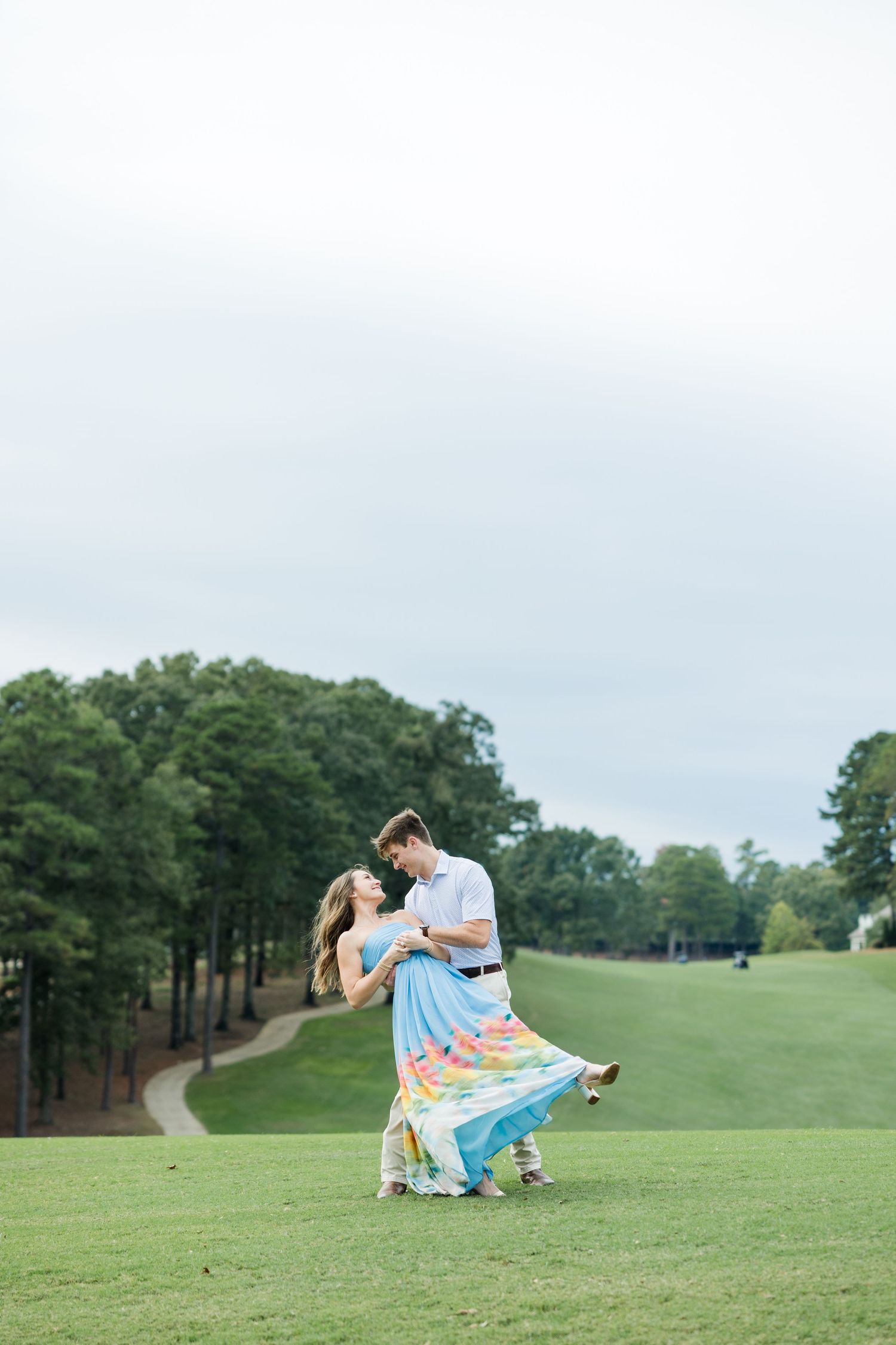 Man dips his fiancée in a romantic dance on a grassy hill surrounded by tall pine trees during engagement photos.
