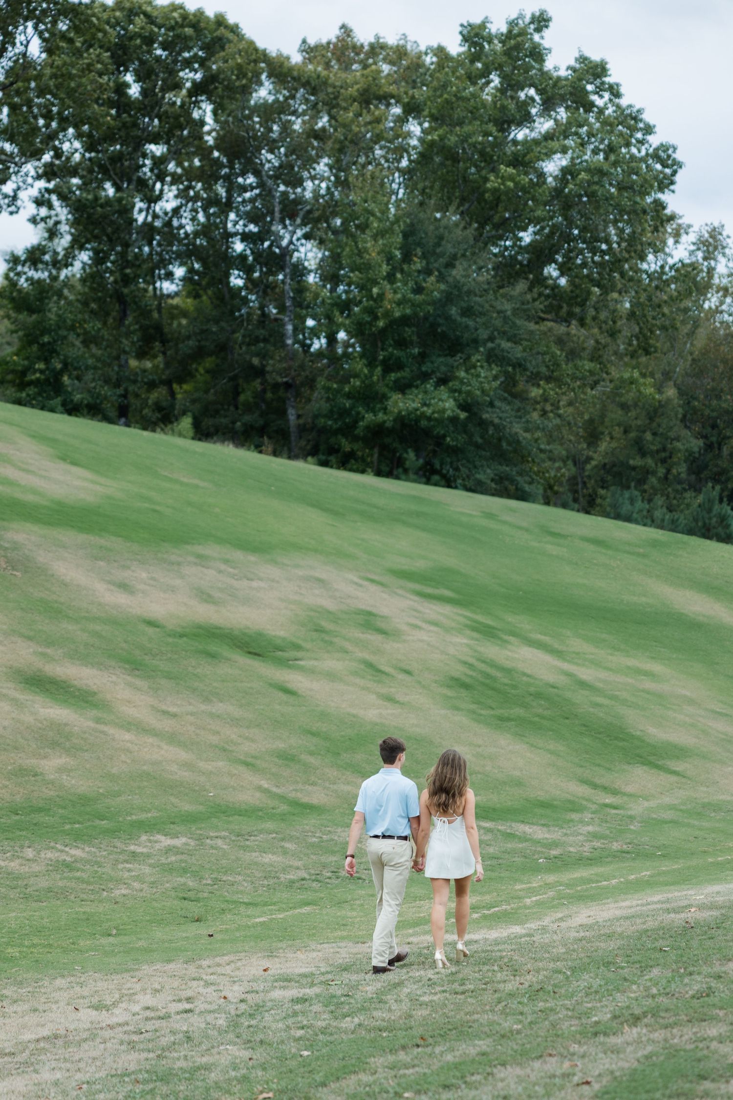 Couple in casual summer outfits walking hand in hand up a grassy hillside toward a line of tall trees.