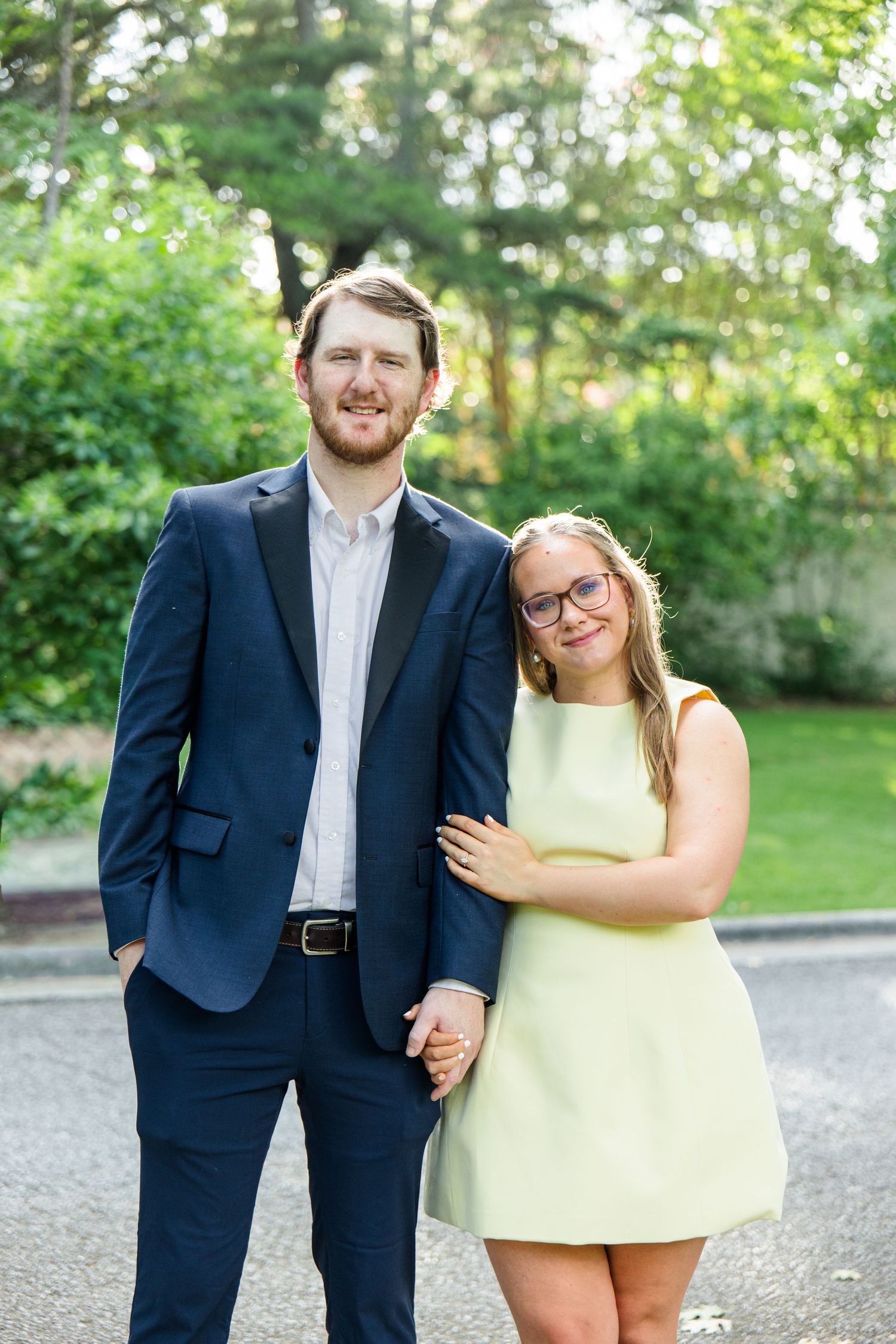 Couple posing outdoors, man in navy suit, woman in light green mini dress with glasses, surrounded by lush greenery.